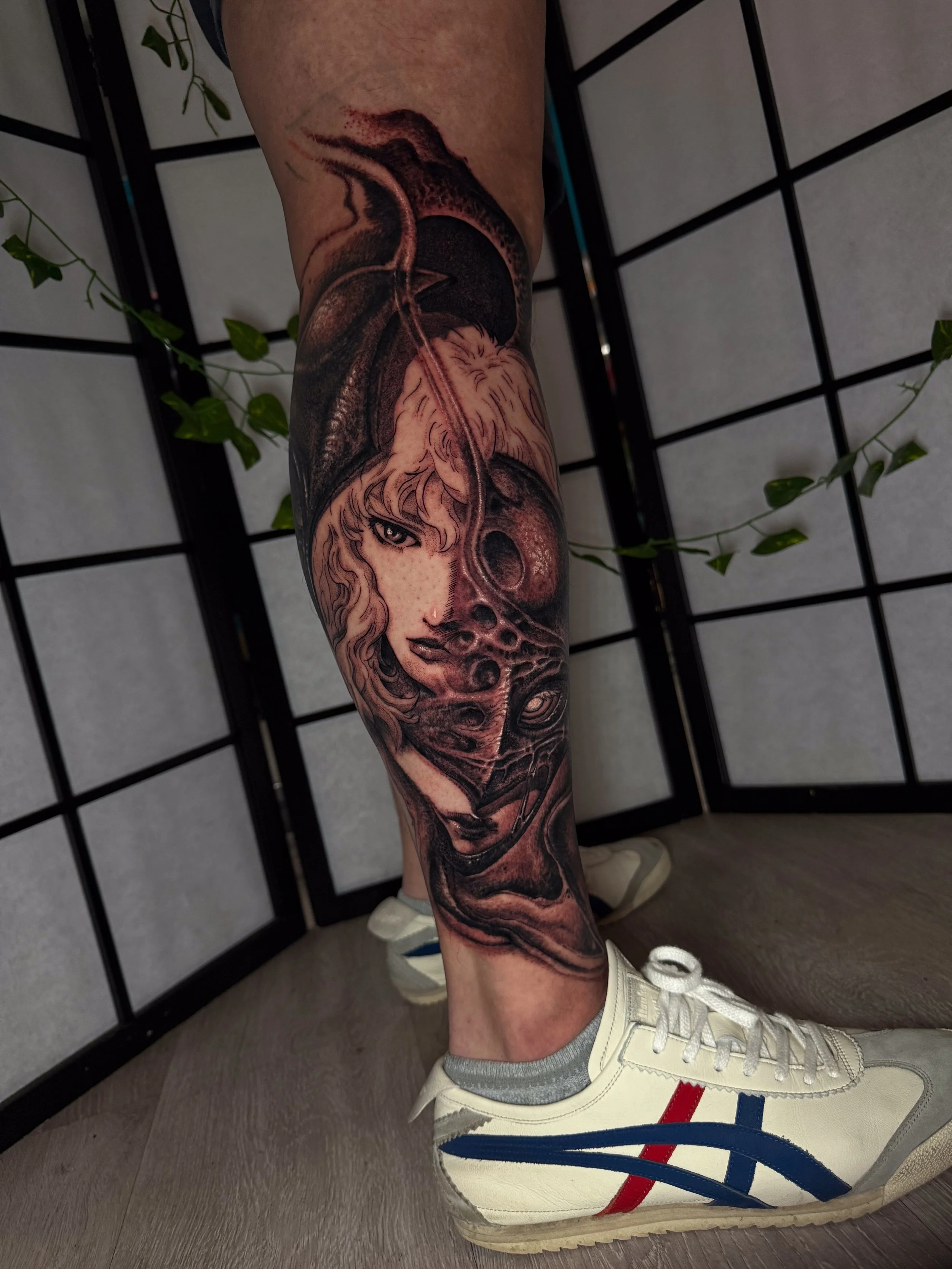 A person with a detailed black and gray tattoo sleeve on their arm, featuring a woman’s face and a wolf, wearing a hood. They are standing in front of a black grid divider with green vines, wearing beige sneakers and white sneakers with red and blue 