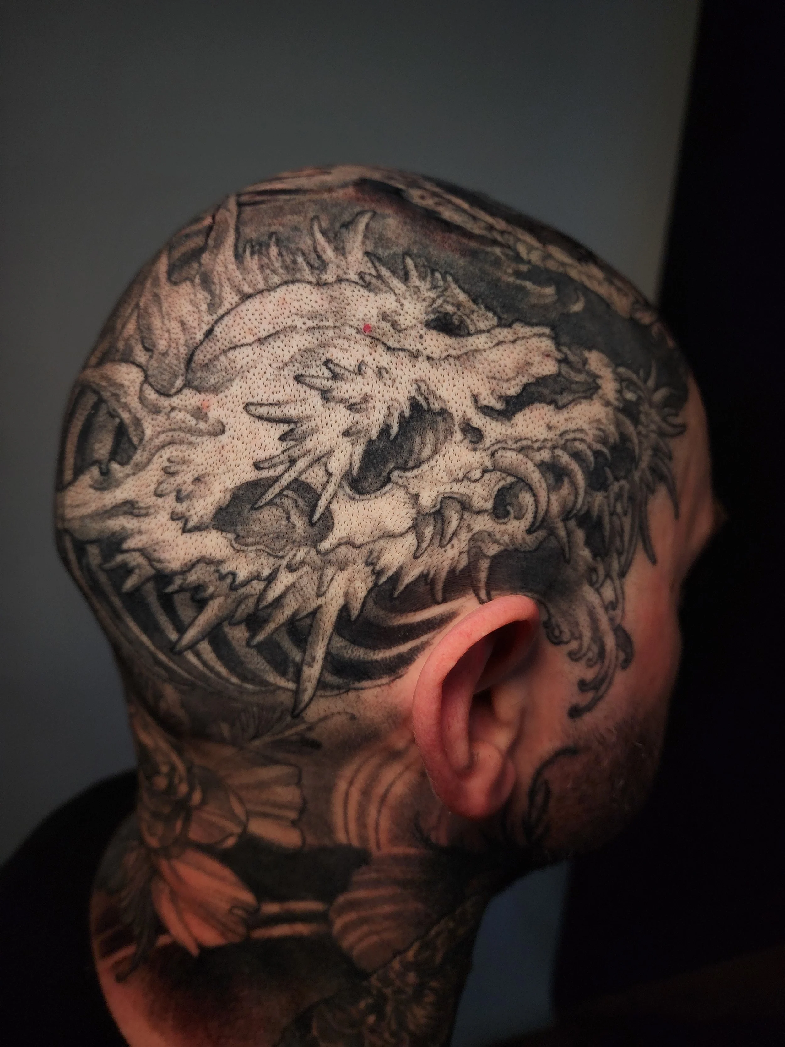 Close-up of a person's head with black and gray tattoo art covering the scalp, neck, and shoulder, featuring an intricate dragon design.
