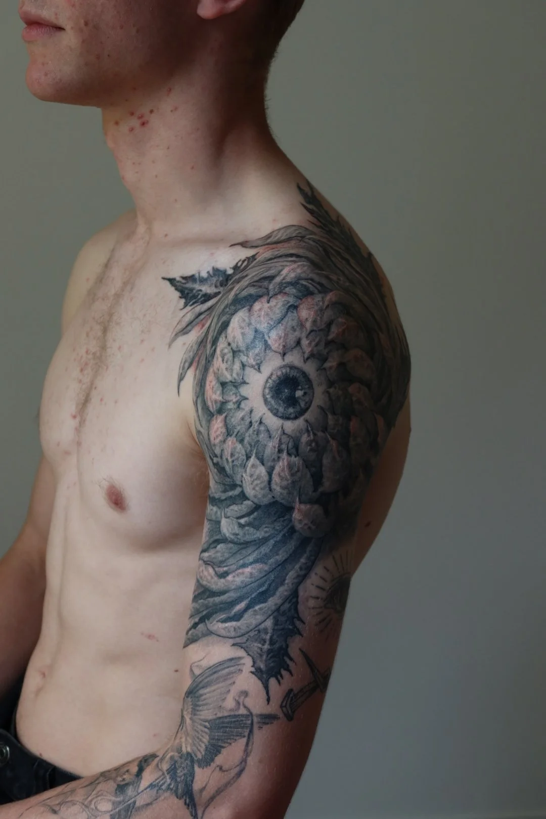 A shirtless man with tattoos on his left arm and shoulder, including a detailed eye within a sunflower and a flying fish tattoo, against a plain background.