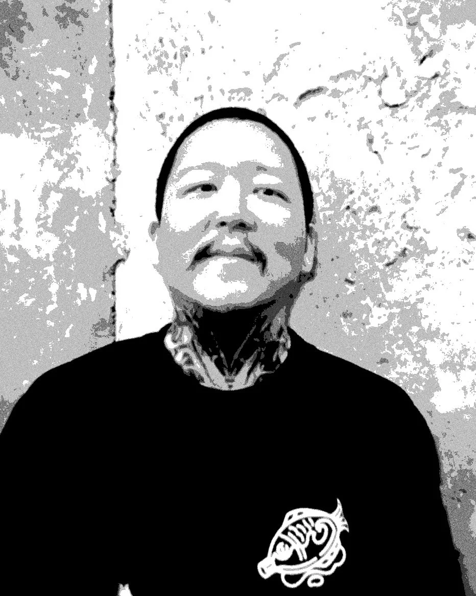 A person with tattoos on their neck and face, wearing a black shirt with a logo, standing against a textured wall.
