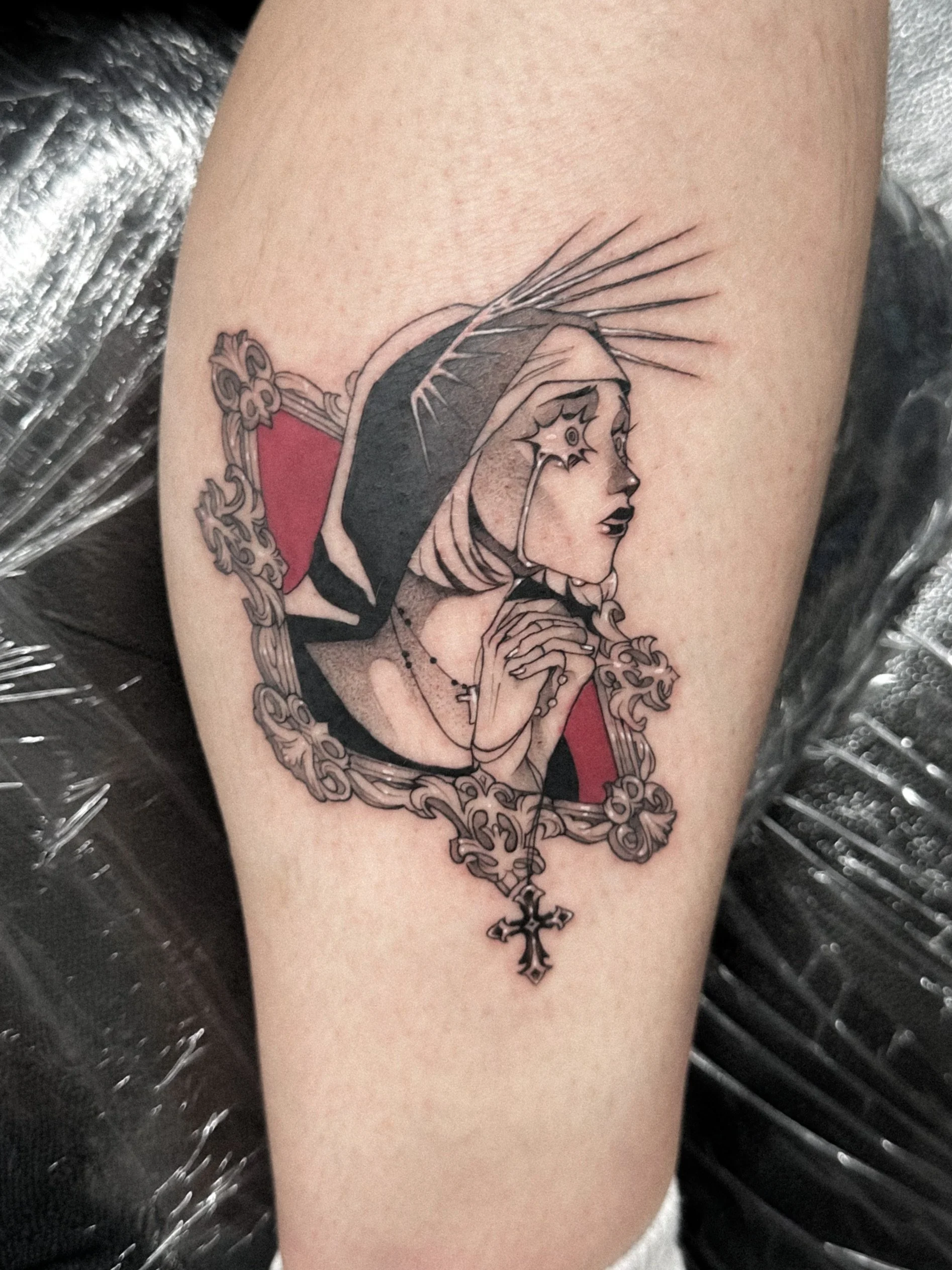 Tattoo of a woman with a headdress and profile view, surrounded by ornate frame, with hands clasped near her face, black and gray ink with red accents.