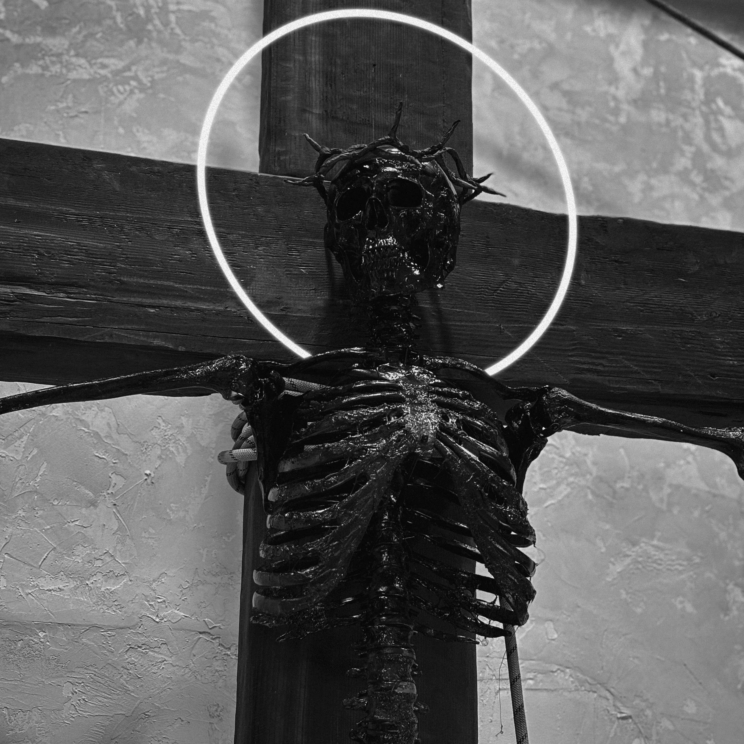 Black and white photo of a skeletal figure with a skull, ribs, and arms, mounted on a wooden cross, with a glowing ring around its head.