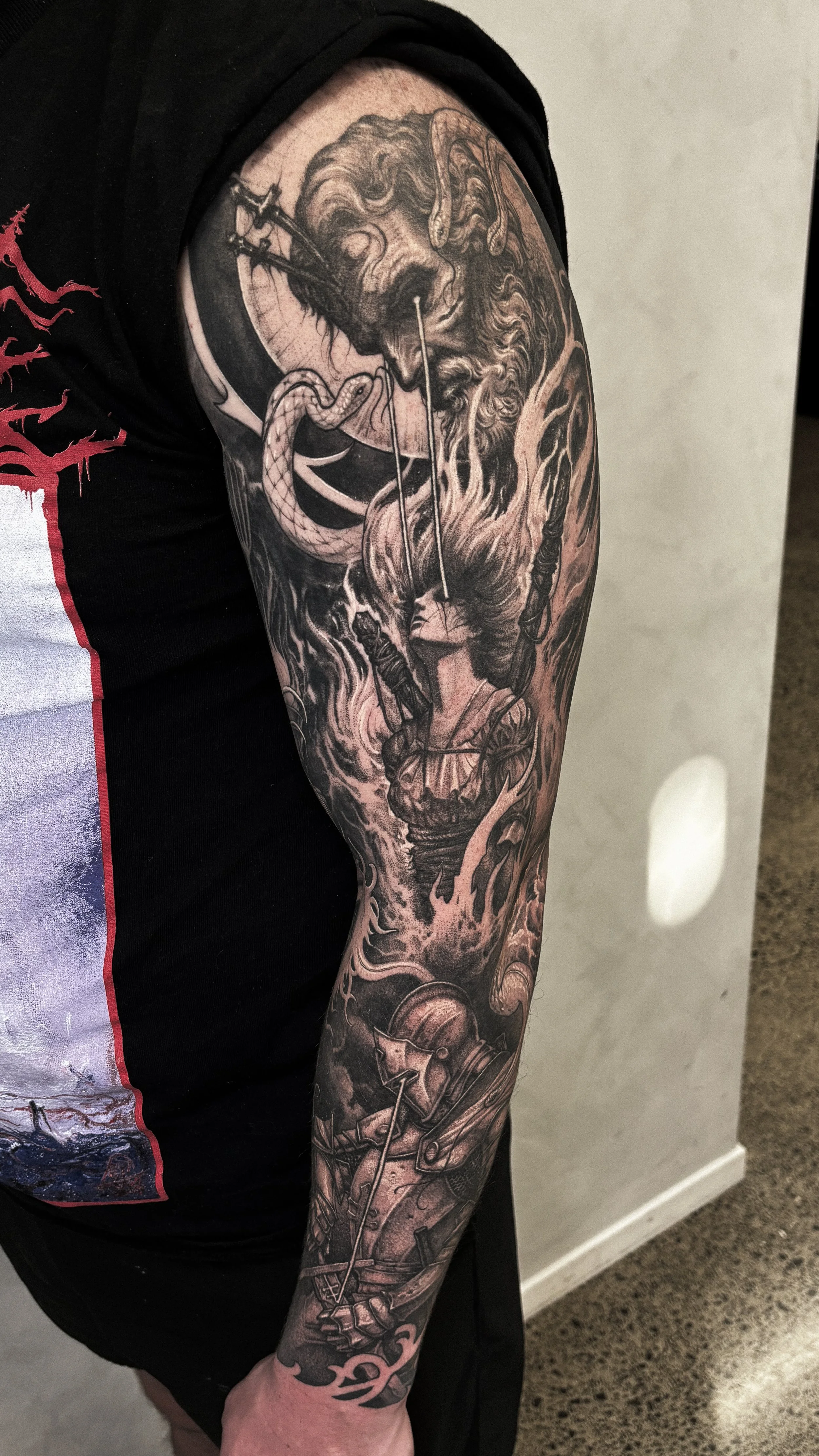 A detailed black and gray sleeve tattoo featuring a lion's head, a snake, a knight with a shield, and a knight in armor holding a sword, with swirling clouds and flames.