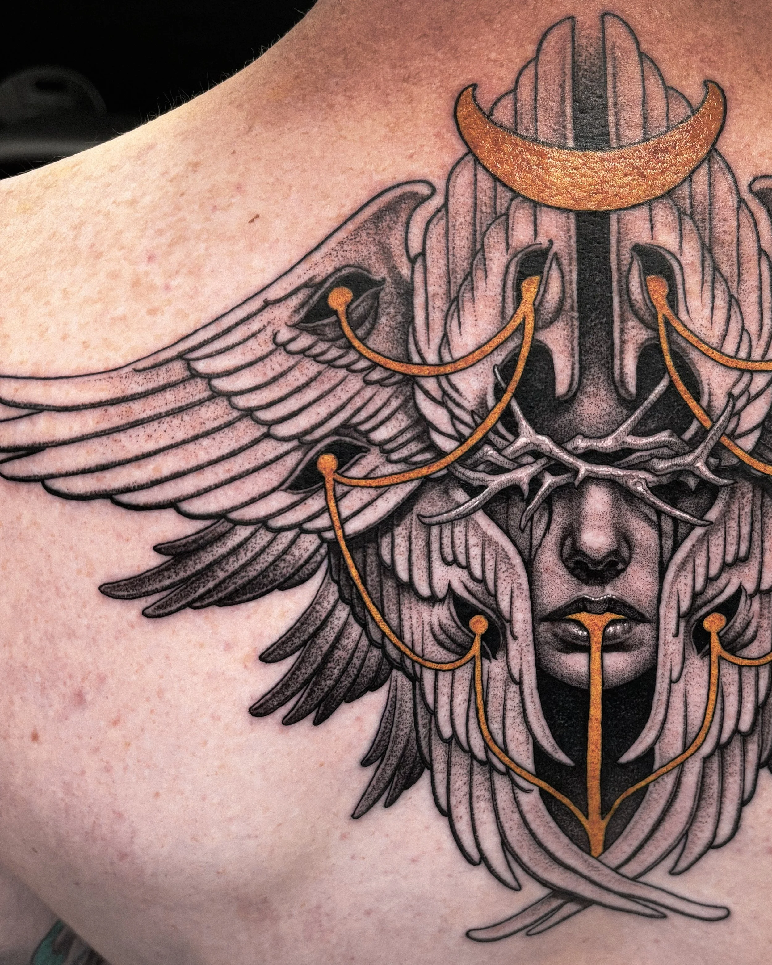Tattoo of a woman's face with wings, a crescent moon, and gold decorative elements.