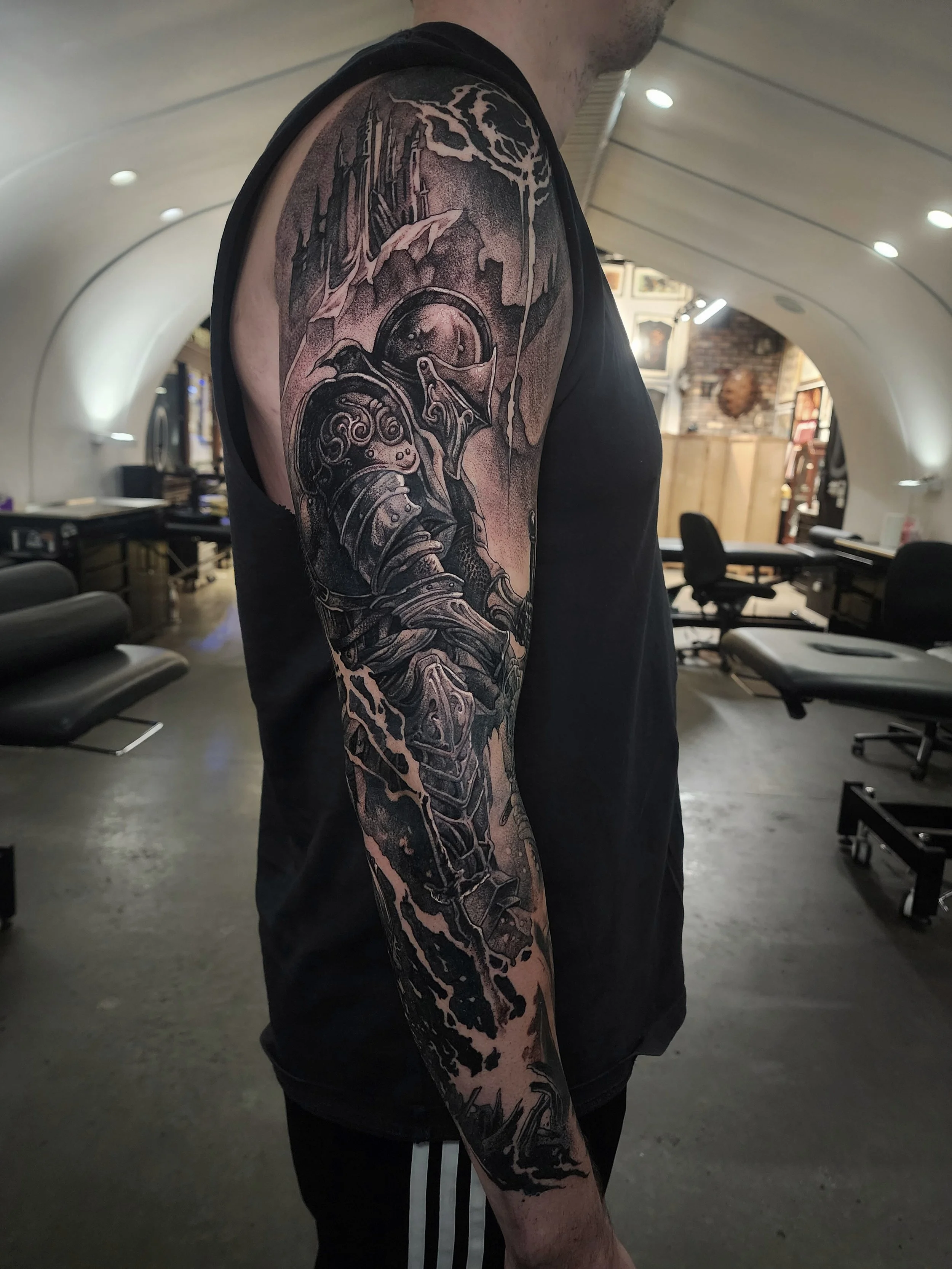 Tattoo of a futuristic soldier in armor on a man's upper arm, with a sci-fi landscape and an abstract sky in black and gray ink.