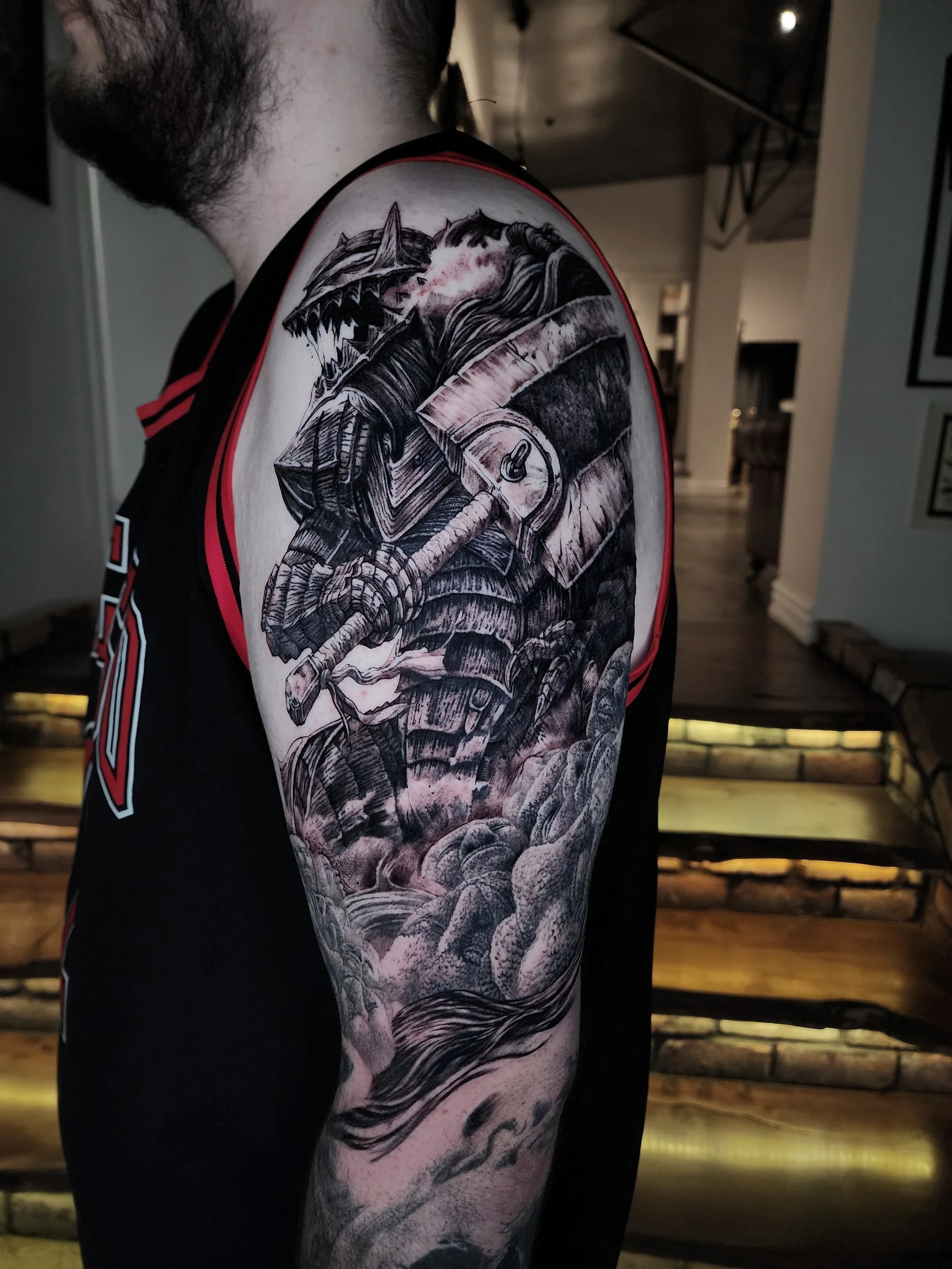 A close-up of a person's upper arm tattoo featuring a detailed black and gray dragon with sharp teeth, horns, and scales, holding a sword. The background includes clouds and mountainous elements.