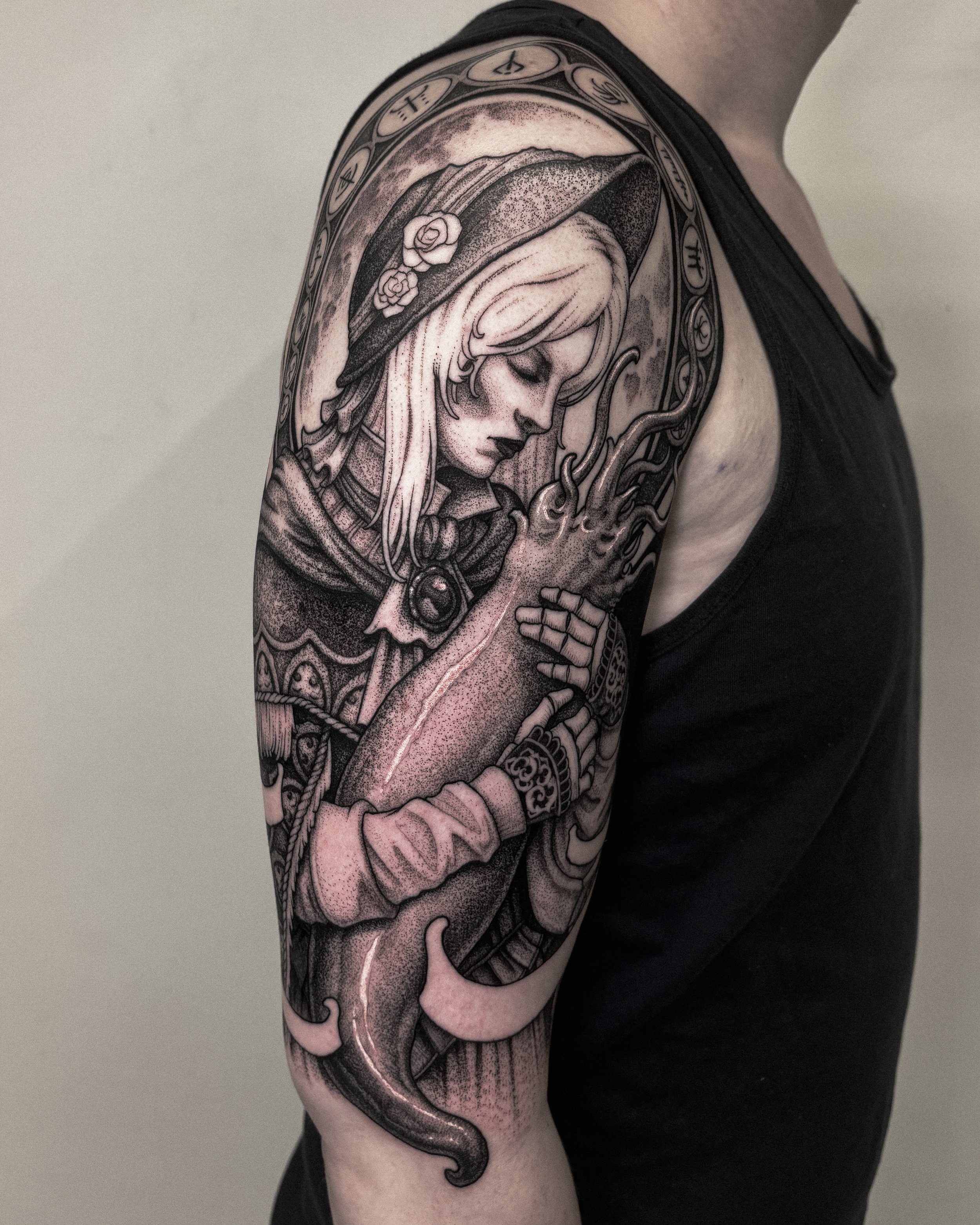Black and gray tattoo of a woman with long hair, wearing a hat with flowers, hugging a large squid, with a decorative halo adorned with symbols surrounding her head.