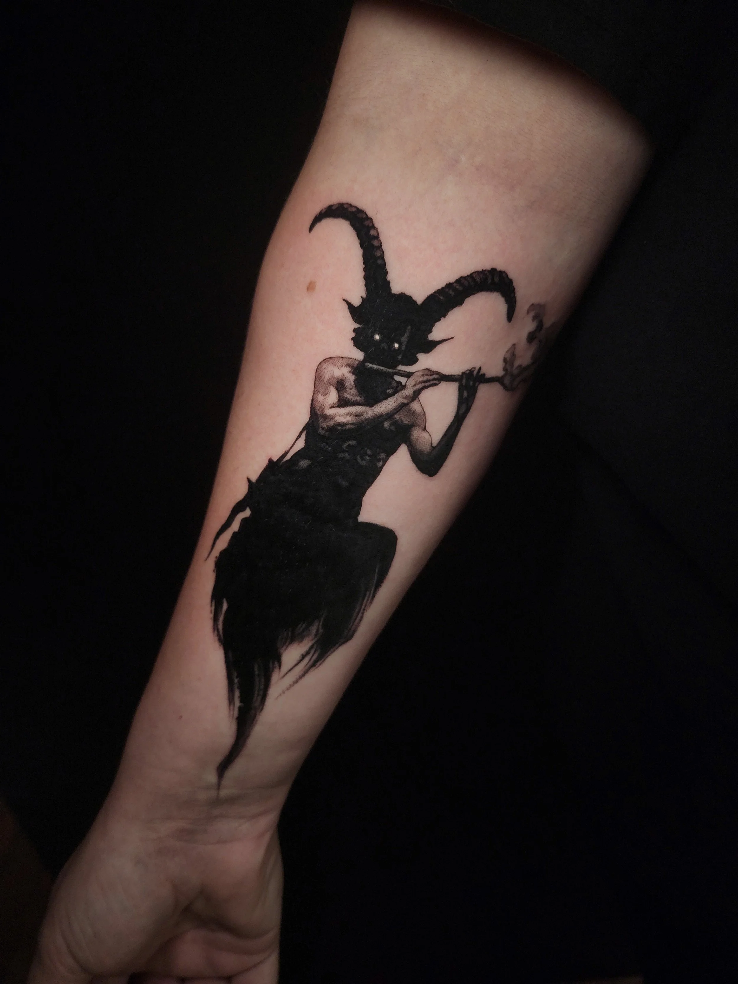 Tattoo of a creature with a goat-like head, long curved horns, and human-like muscular torso playing a flute, with smoke emerging from it.