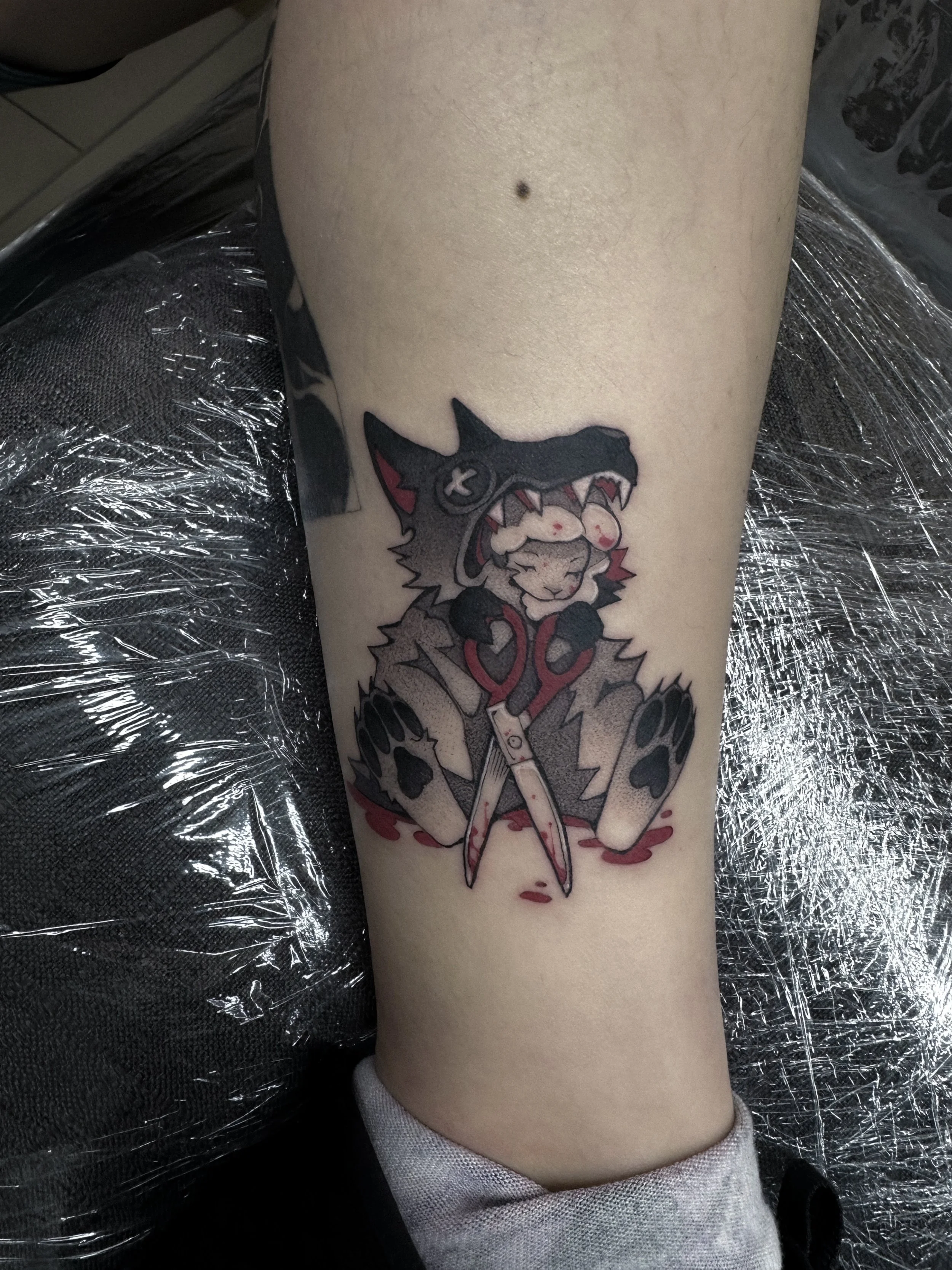 Tattoo of a cartoonish wolf dog character with sharp teeth and red accents on the back of a person's calf, with blood splatters around the tattoo.