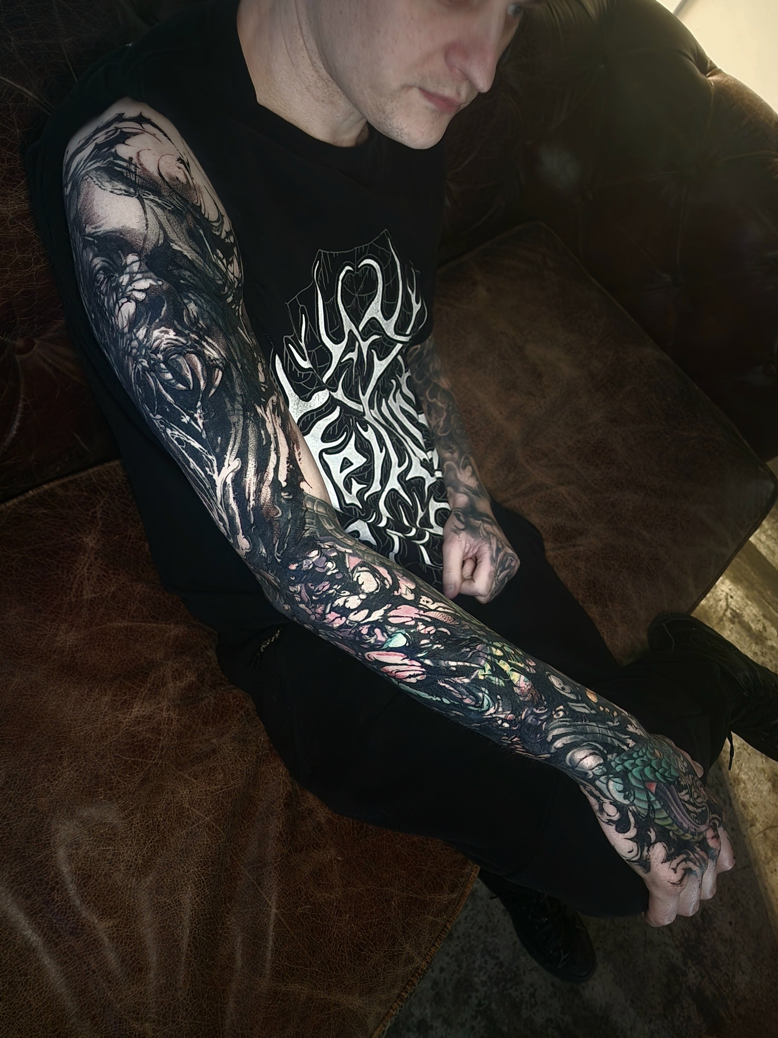 A man with extensive black and colorful tattoo sleeves on his arms, sitting on a brown leather couch, wearing a black T-shirt with a graphic design. The photo is taken from above, capturing his partly visible face and arms.