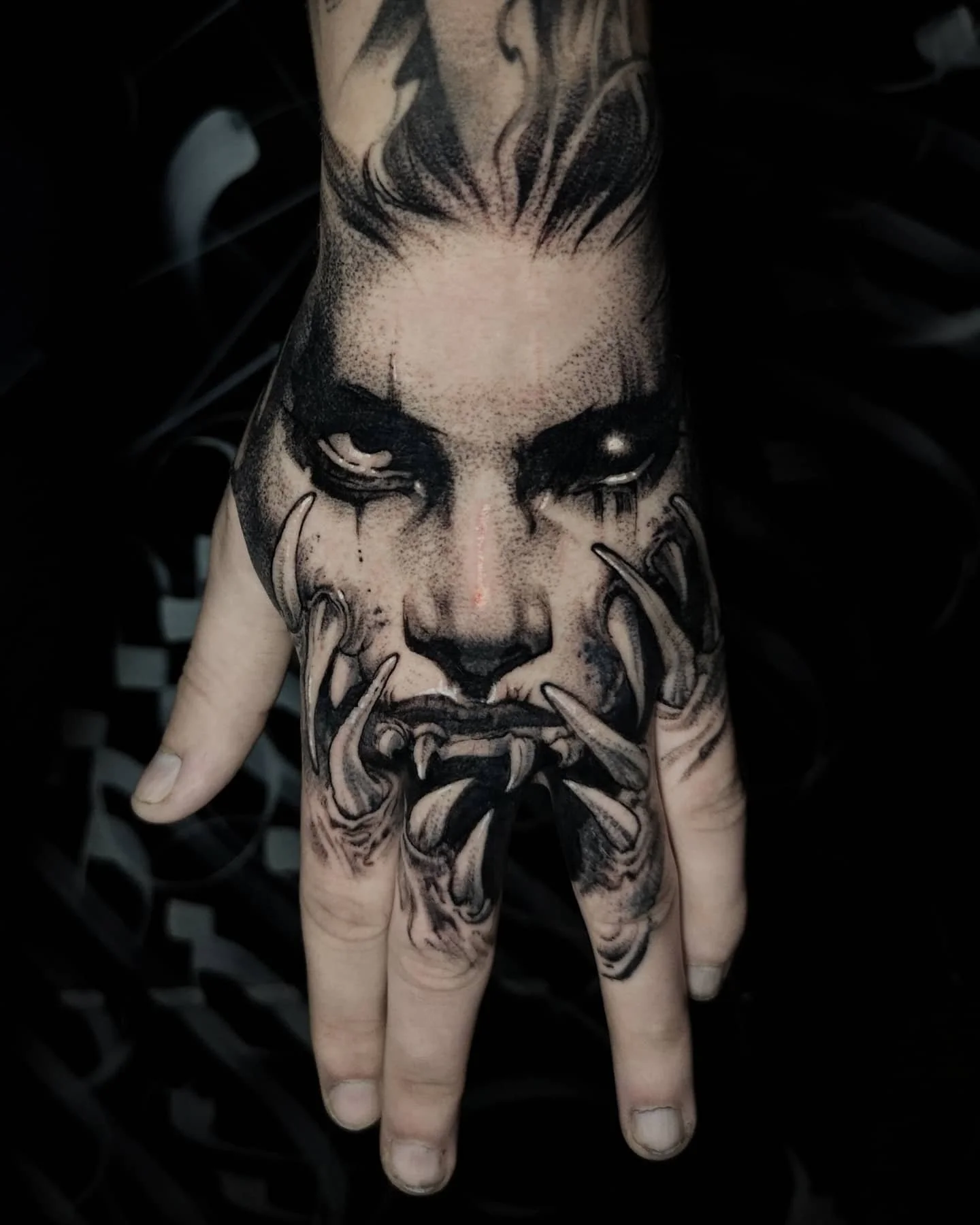 A tattooed hand with black and gray tattoo of a face with an intense expression, featuring black eyes with tears, a fierce face with large teeth, and tentacle-like elements extending from the lower face.
