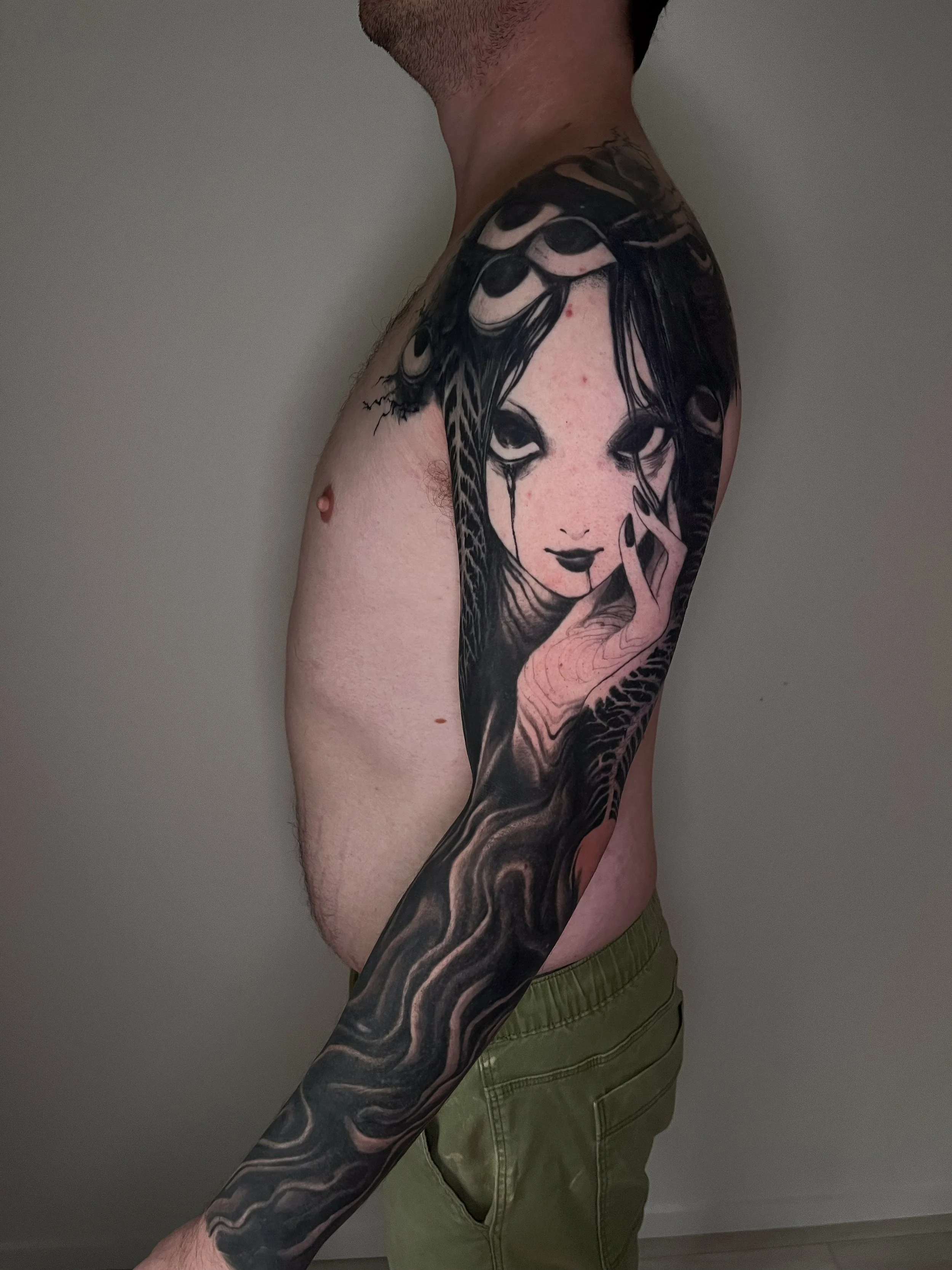 A person with a large, detailed black and gray tattoo of a woman's face and hands with long hair that appears to blend into a dragon or snake pattern, covering the shoulder and arm.