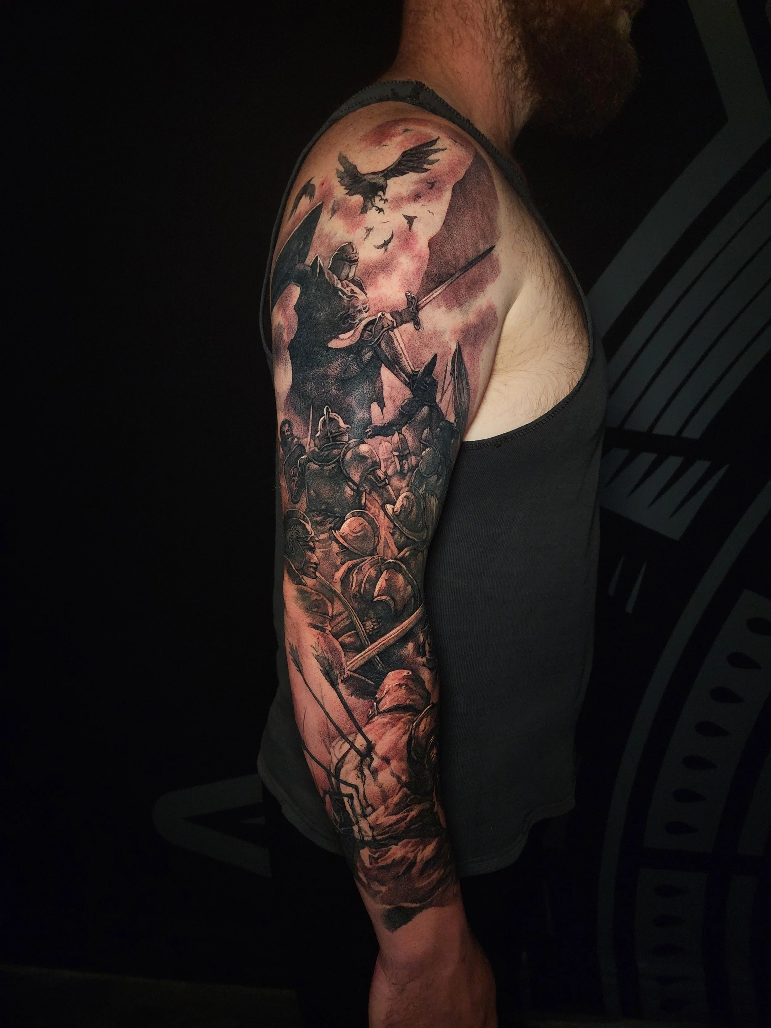 A detailed black and gray tattoo sleeve depicting a medieval battle scene with armored soldiers, a knight with a sword, a dragon, and a flock of birds against a dark sky.