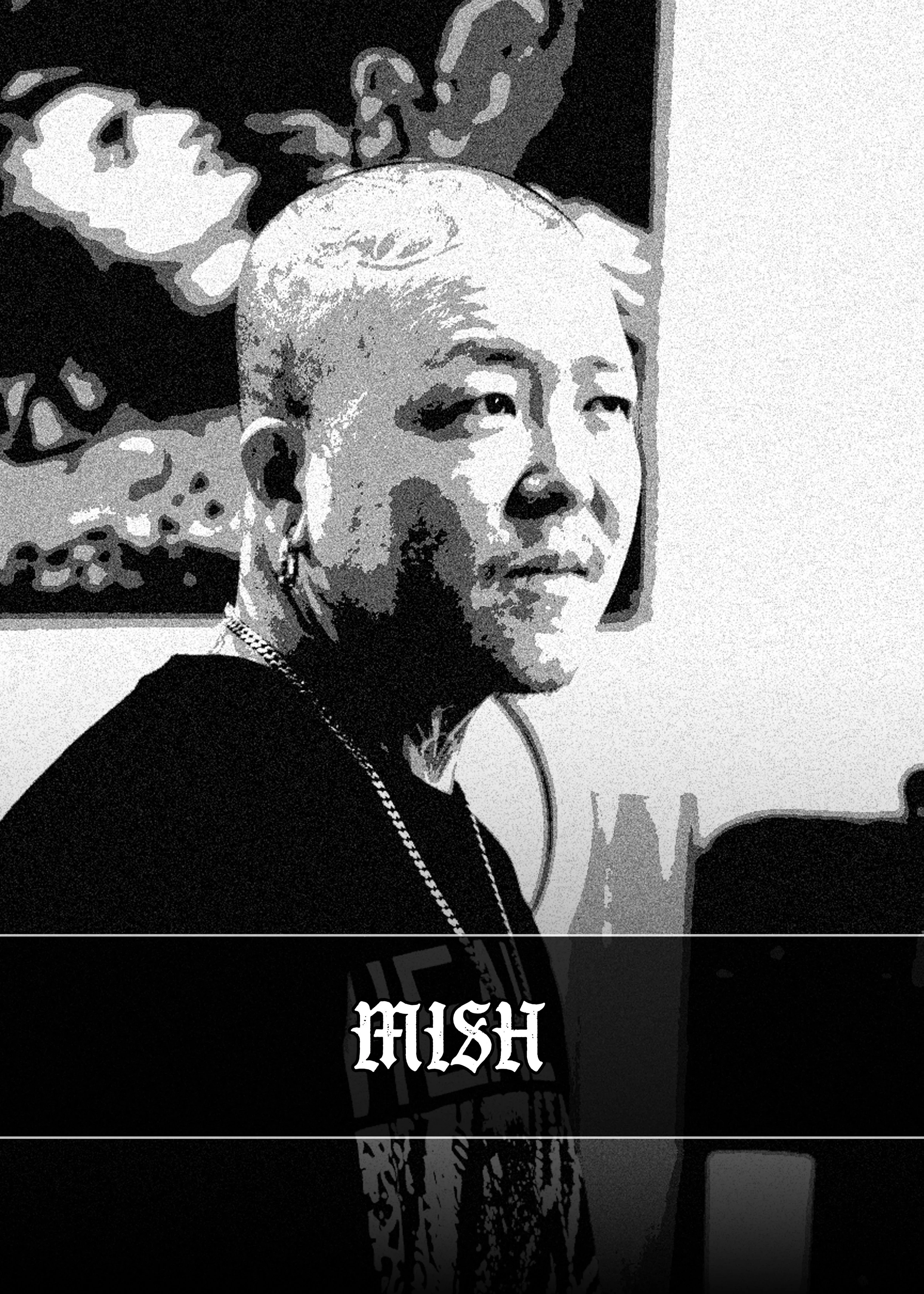 A black and white portrait of a woman with short, platinum blonde hair, wearing earrings, a chain necklace, and a black shirt with the word 'MISH' printed on it, standing in front of a framed artwork.