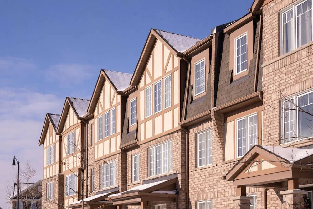 A row of multi-story townhouses with brick and beige painted wood siding, white-framed windows, and steep gabled roofs under a clear blue sky.