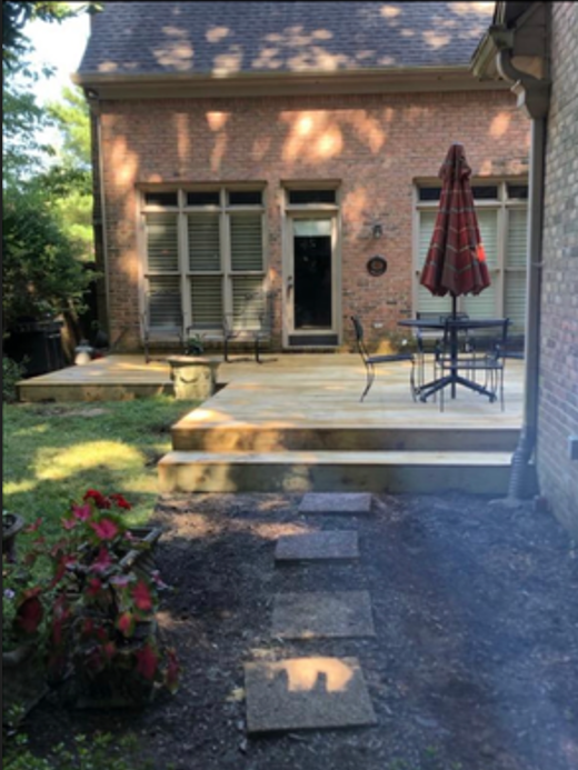 Backyard patio with steps leading up to a brick house with large windows and a door, outdoor table with a closed red umbrella, and some garden plants.
