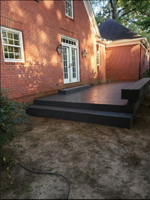 Newly built black wooden deck with steps attached to a brick house with white-framed windows and a white door with glass panels, located in a backyard with some greenery and dirt ground.