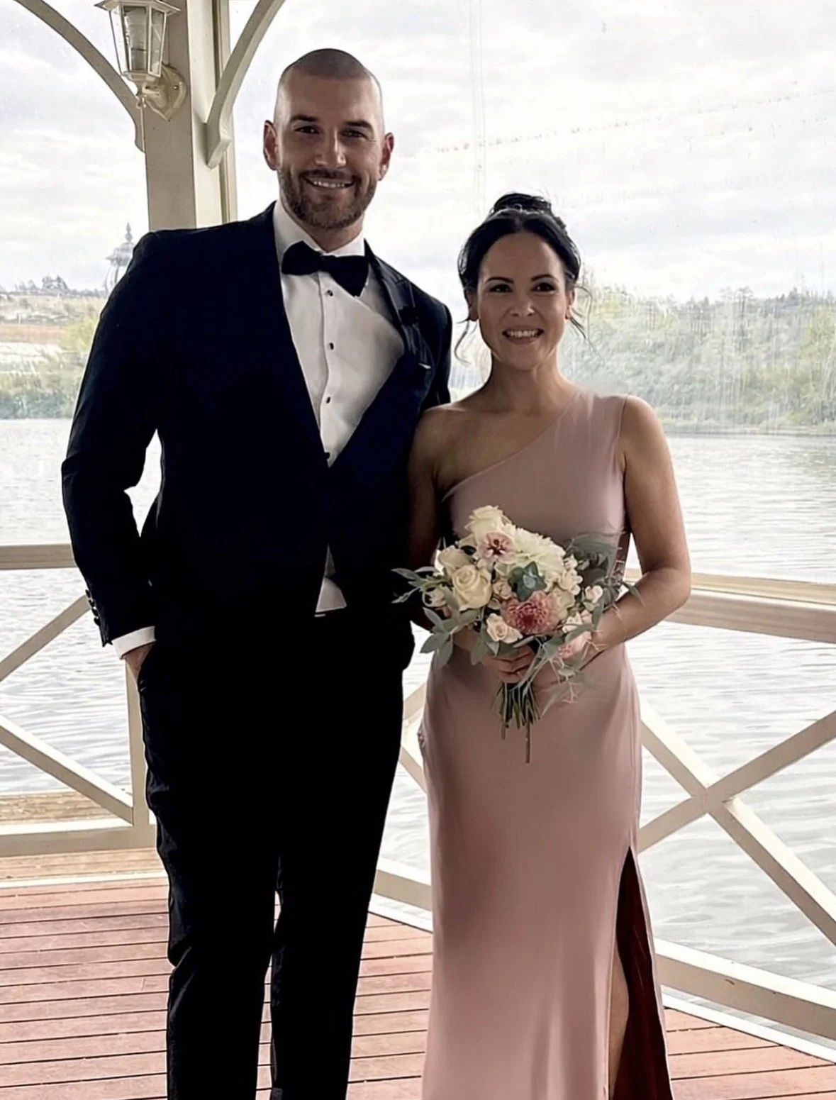 A man in a black tuxedo and a woman in a pink dress holding a bouquet of flowers, standing on a boat deck with water and trees in the background.