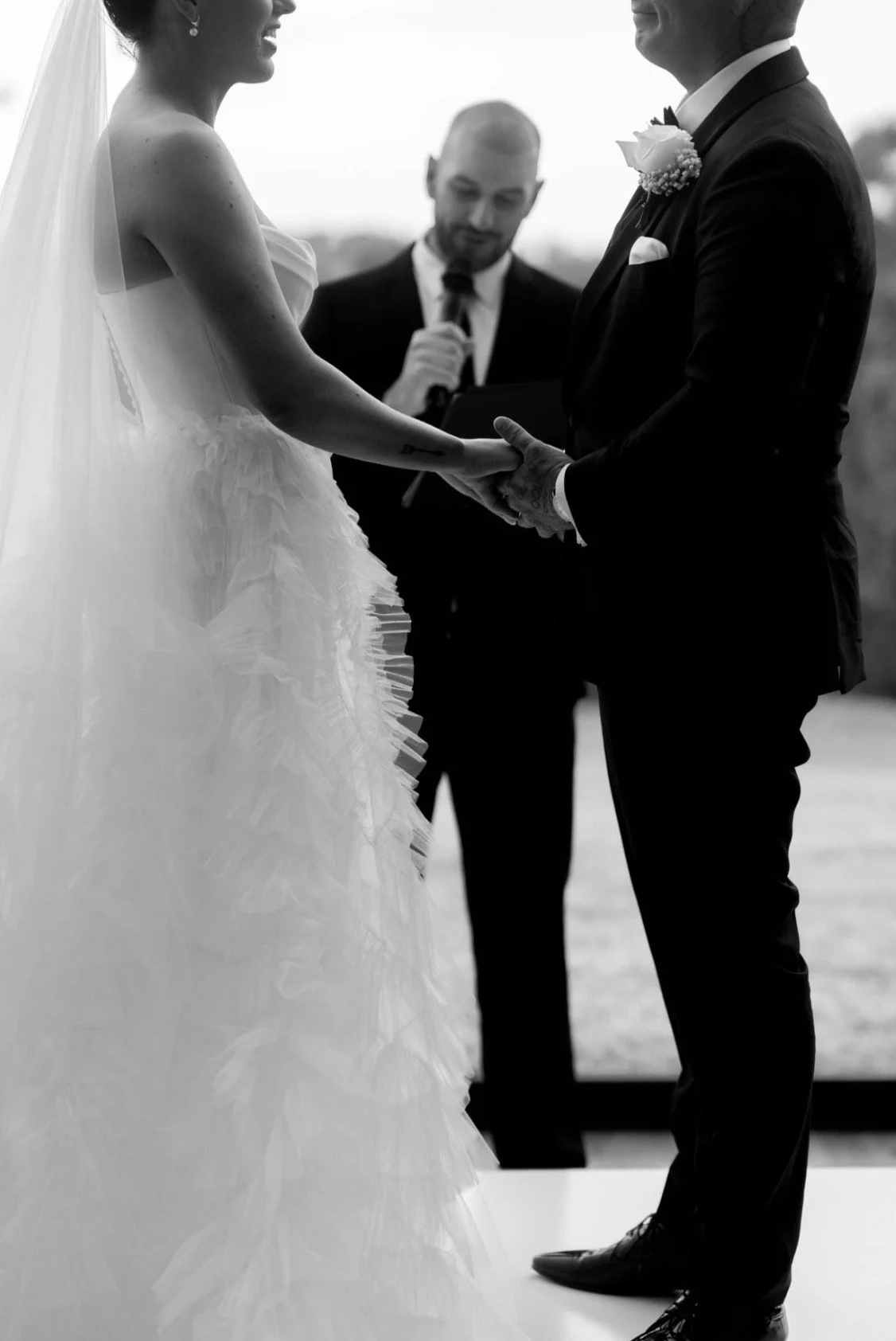 A black-and-white photo of a bride and groom holding hands during their wedding ceremony, with an officiant standing behind them reading from a book.