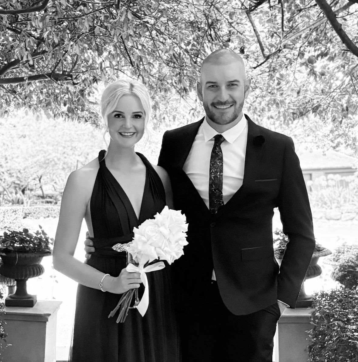 A black and white photo of a smiling man and woman dressed in formal attire outside. The woman holds a bouquet of flowers. They are standing in front of trees and garden decorations.