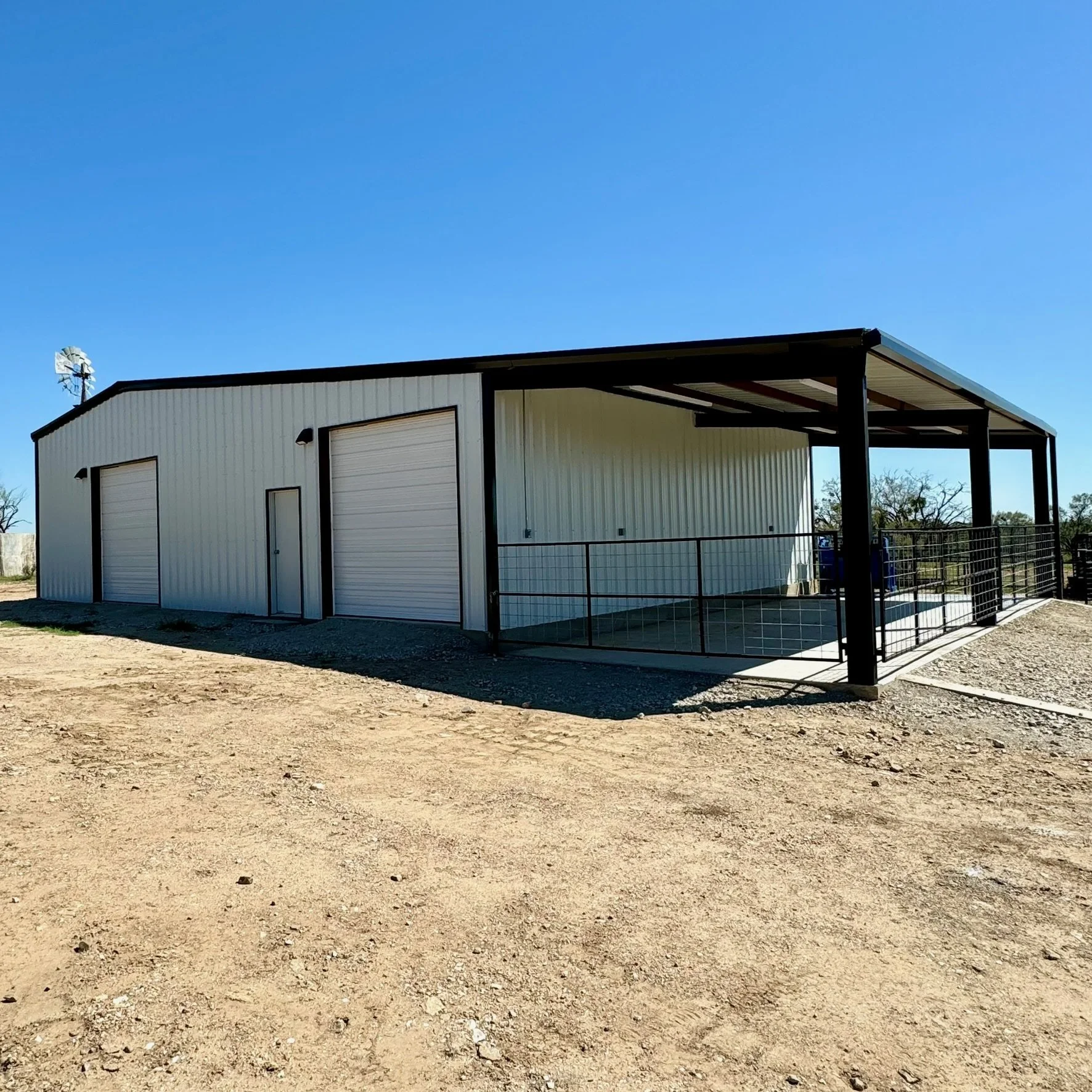 A large metal building with two roll-up doors, a small personnel door, and a covered outdoor area with a metal railing, set in a dirt lot under a clear blue sky.