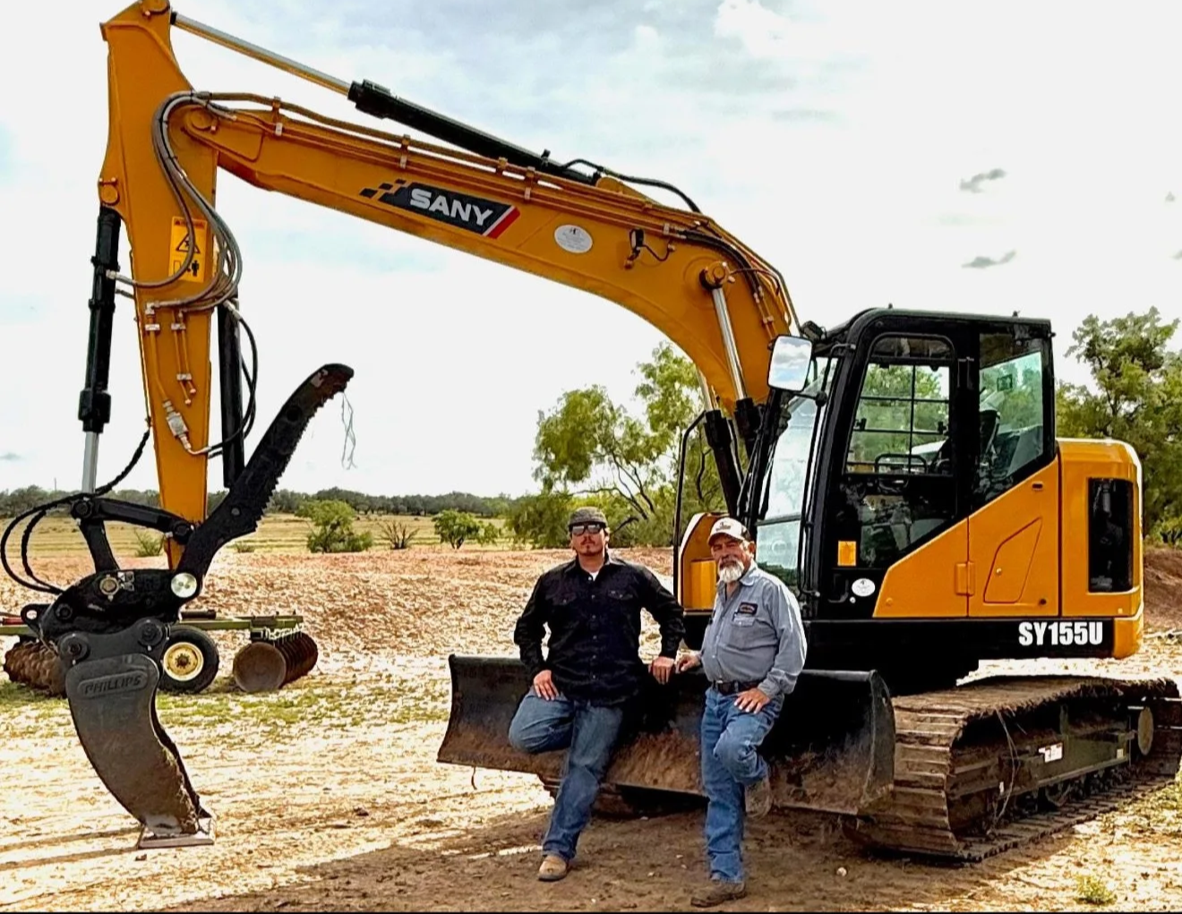 Two men in work clothes and caps posing next to a yellow SANY excavator in a rural area with sparse trees and an open field.