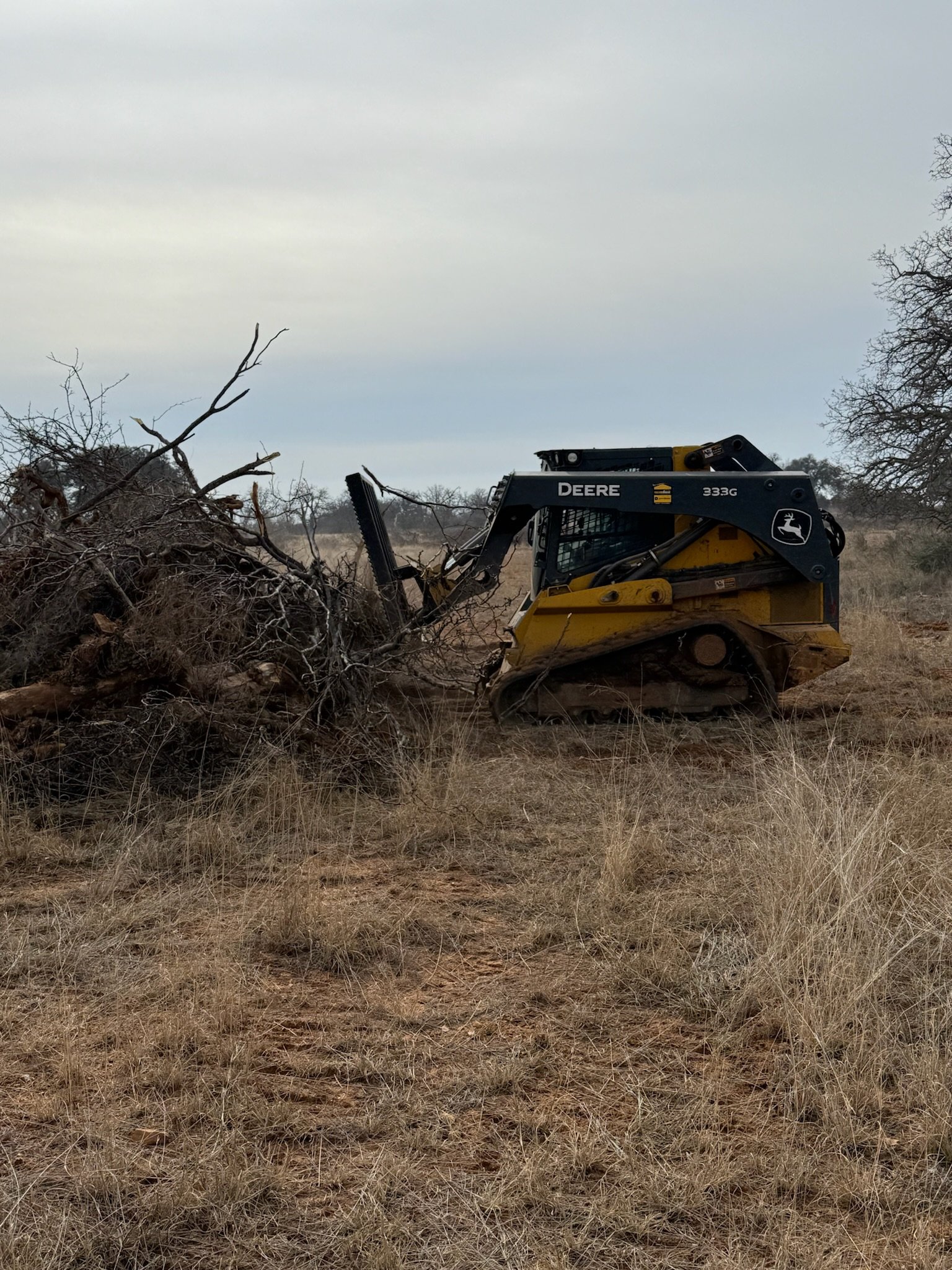 A yellow and black John Deere 333G compact track loader clearing dry, tangled branches and brush in a field with tall grass and a cloudy sky.