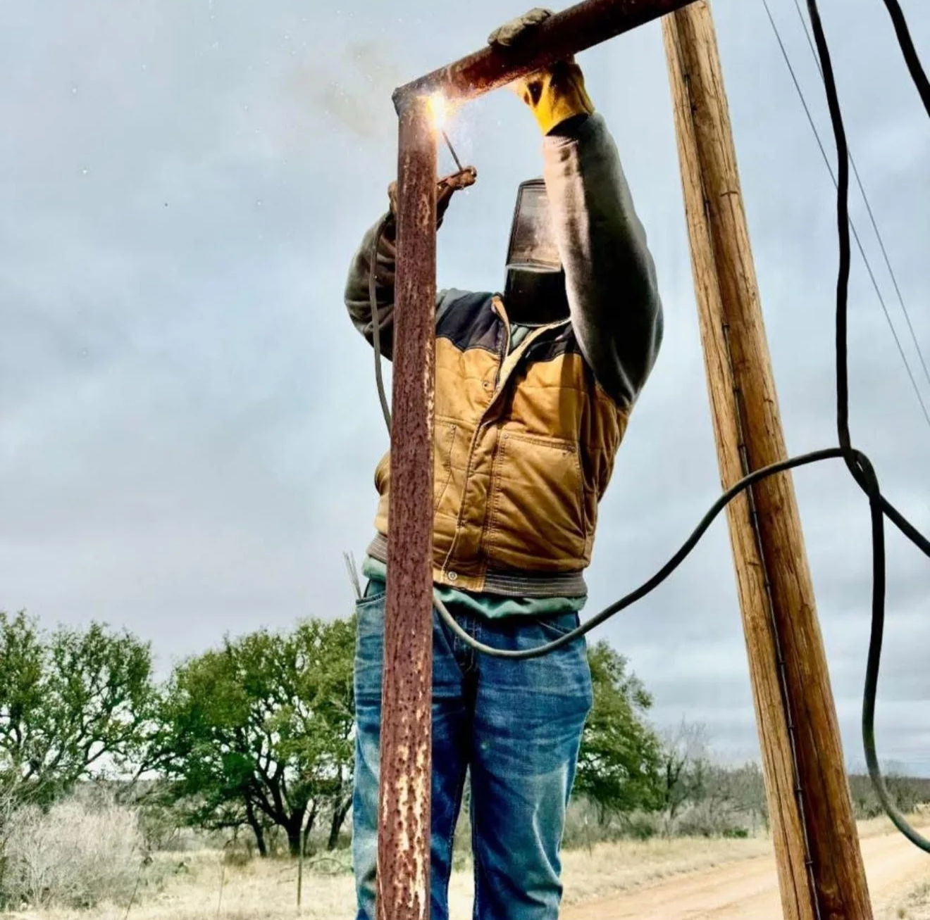 A person wearing a mask and a brown vest welding on a power pole outdoors under a cloudy sky.