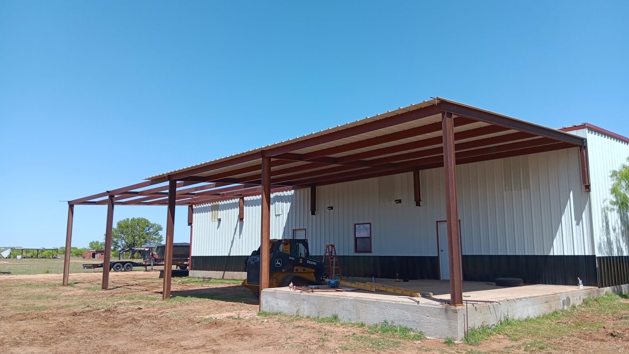 Construction site featuring a partially built metal building with a wooden framework extension, construction tools, a small tractor, and outdoor equipment on a dirt ground under a clear blue sky.