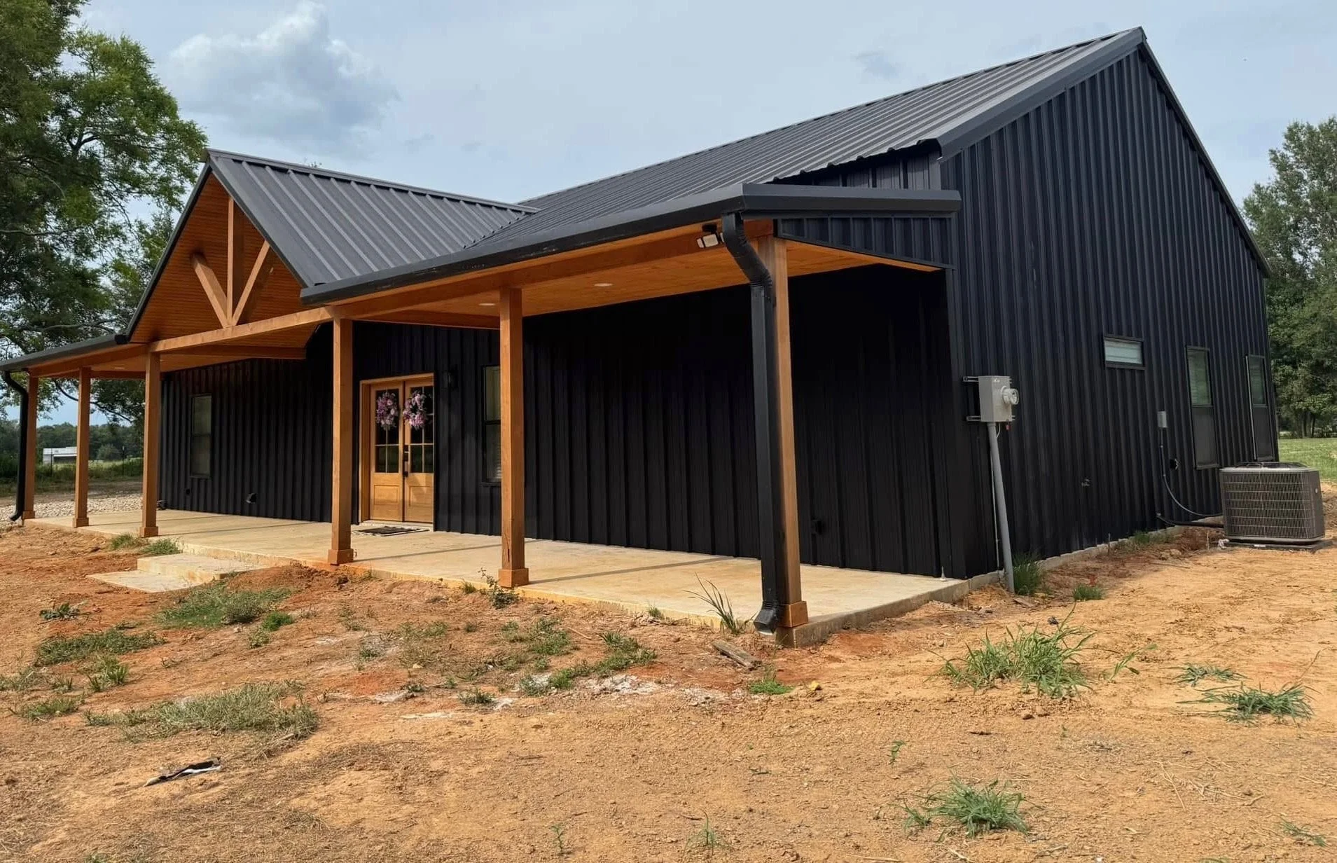 A modern black barn-style house with a wooden front porch and door, on a patch of bare dirt with some grass and a cloudy sky in the background.