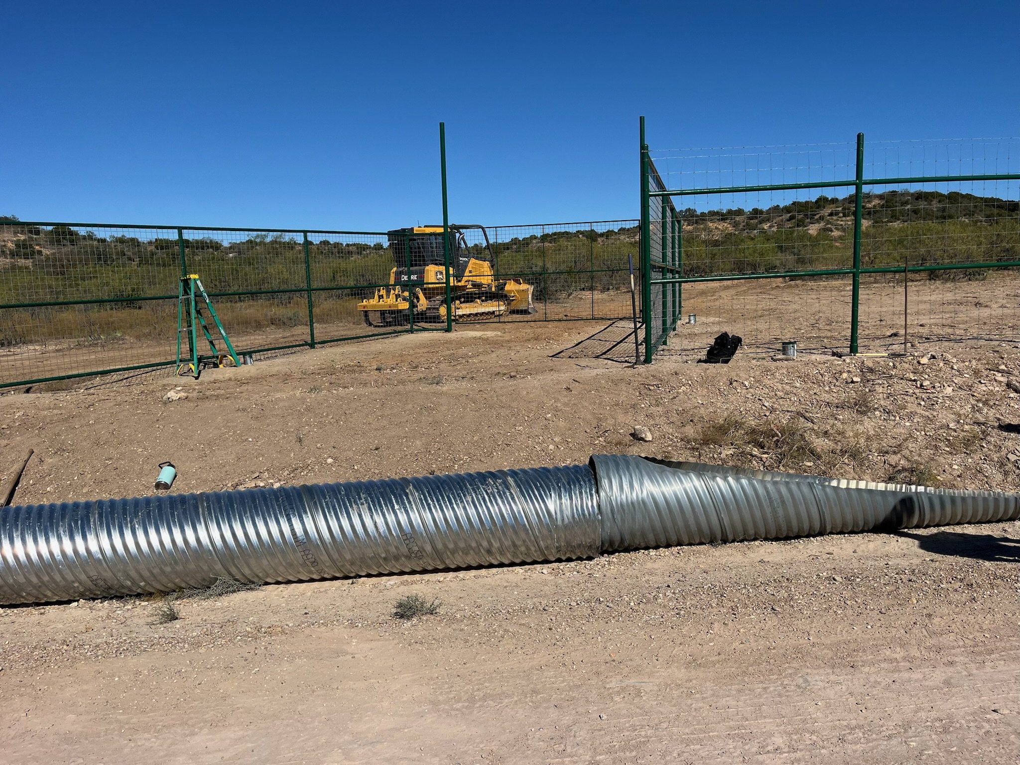 Construction site with green fence, yellow bulldozer, large metal duct lying on dirt ground, and clear blue sky in the background.