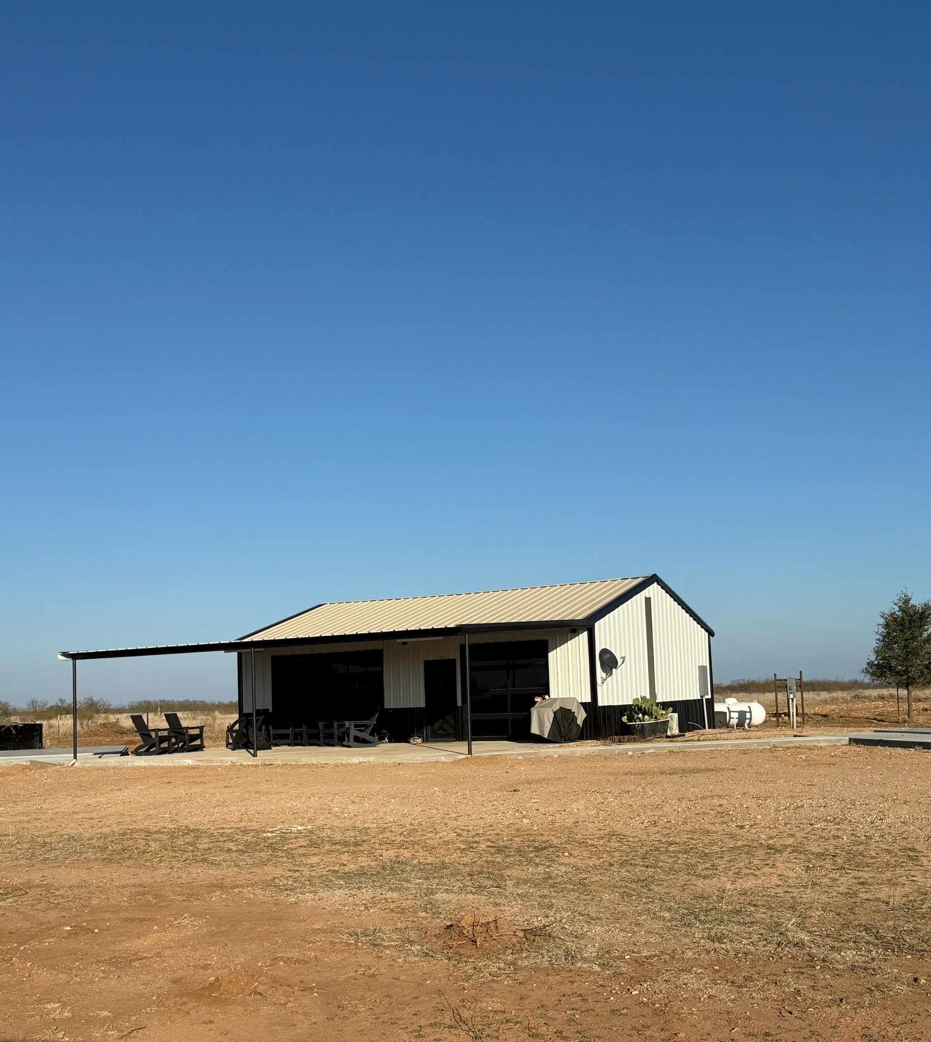 A small, white metal building with a beige roof situated in a rural, flat landscape under a clear blue sky. The building has an attached covered patio with lounge chairs, a barbecue grill, a satellite dish, and potted plants nearby. There are propane