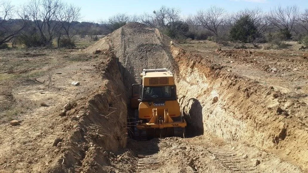 A yellow Deere bulldozer working in a deep trench on a barren construction site with sparse trees in the background.