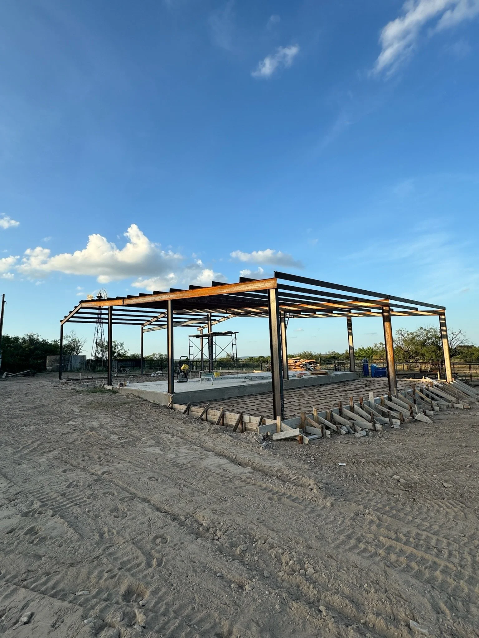 Construction site with a building frame and exposed beams, surrounded by dirt and construction materials under a clear blue sky.