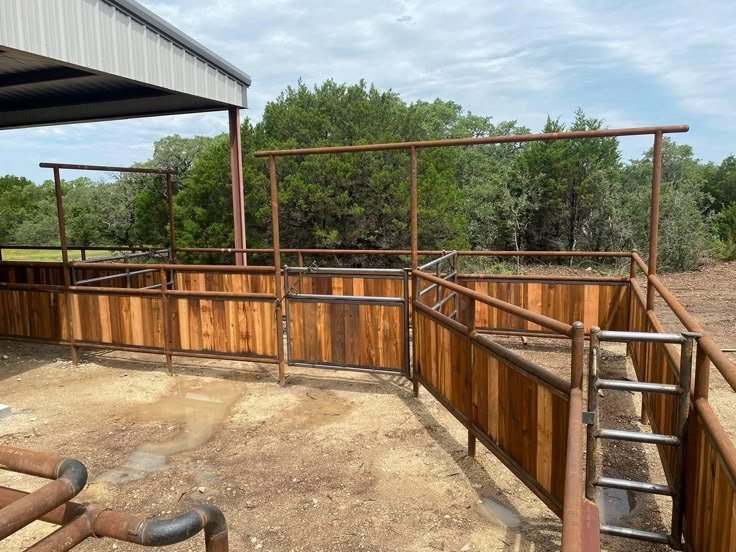 Animal pen with wooden and metal fencing, roller gates, and a dirt ground, set outdoors with trees in the background.