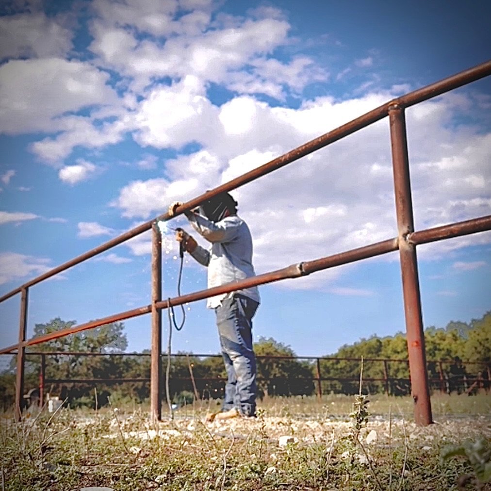A person with dreadlocks and dark skin is standing outdoors on a dirt path, drilling or working on a rusty metal railing under a partly cloudy sky.