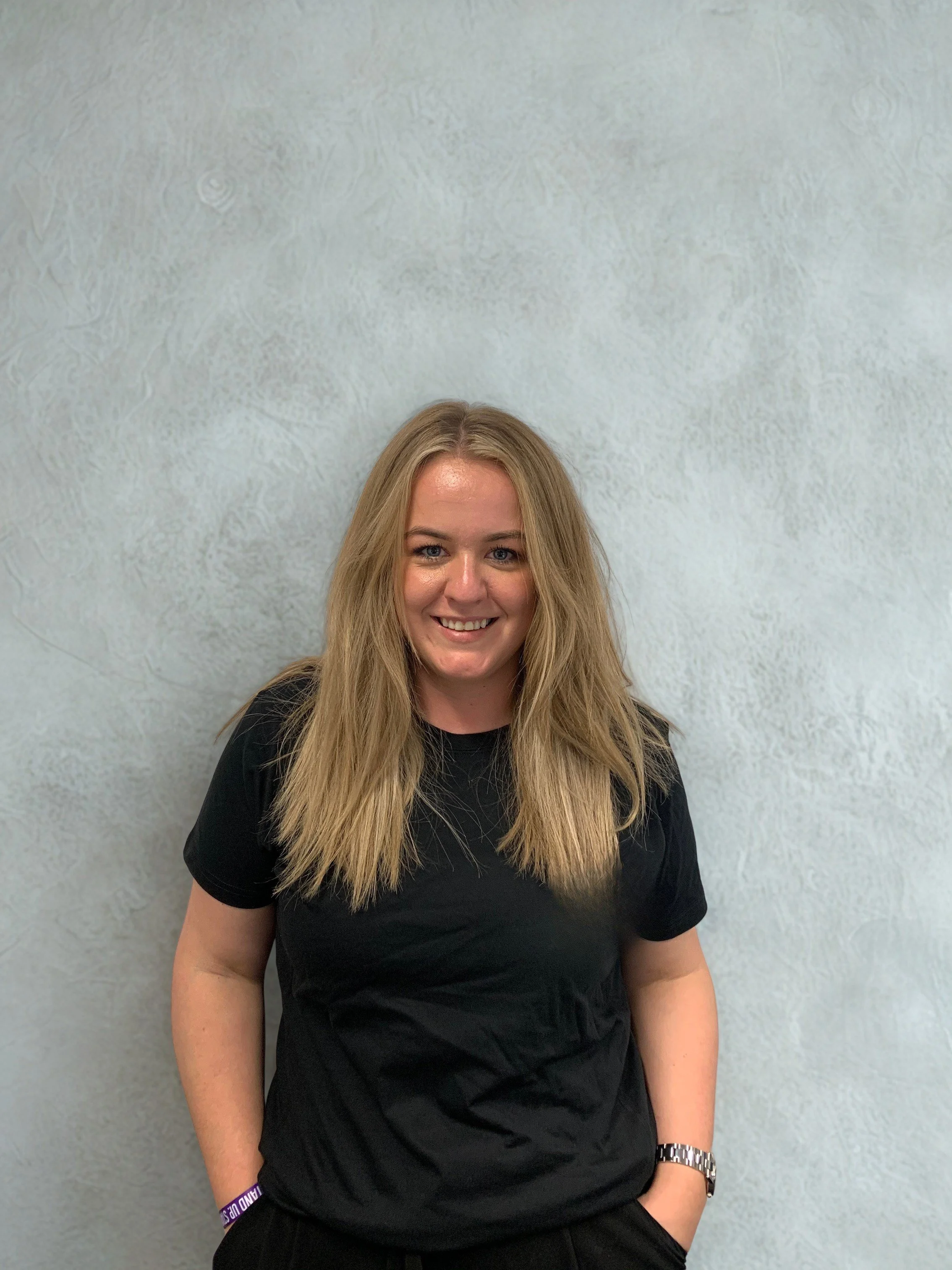A smiling woman with long blonde hair wearing a black t-shirt and a silver watch, standing in front of a textured gray wall.