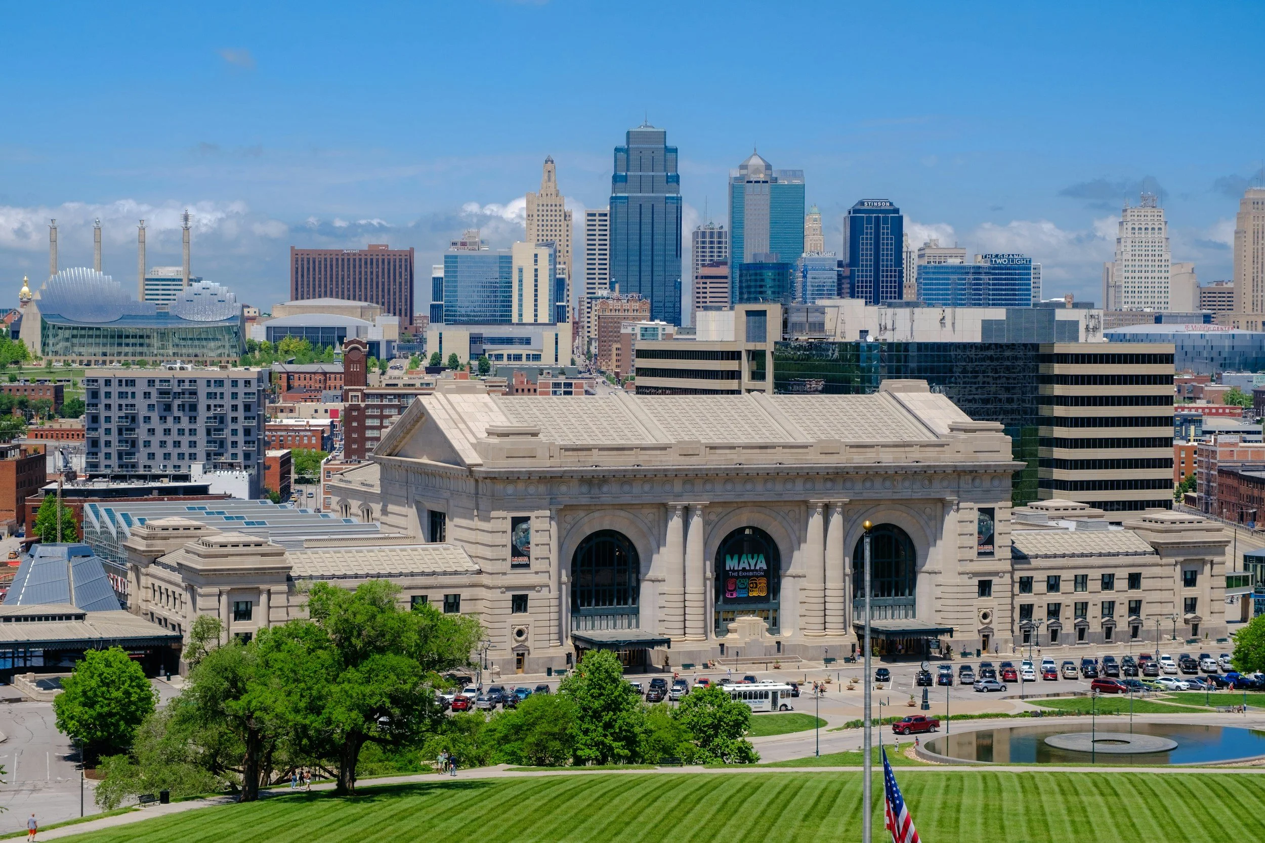 View of downtown Kansas City, Missouri, skyline with tall skyscrapers, a large modern building with a metallic roof, a green park with trees, a pond, and people walking, flags in the foreground.