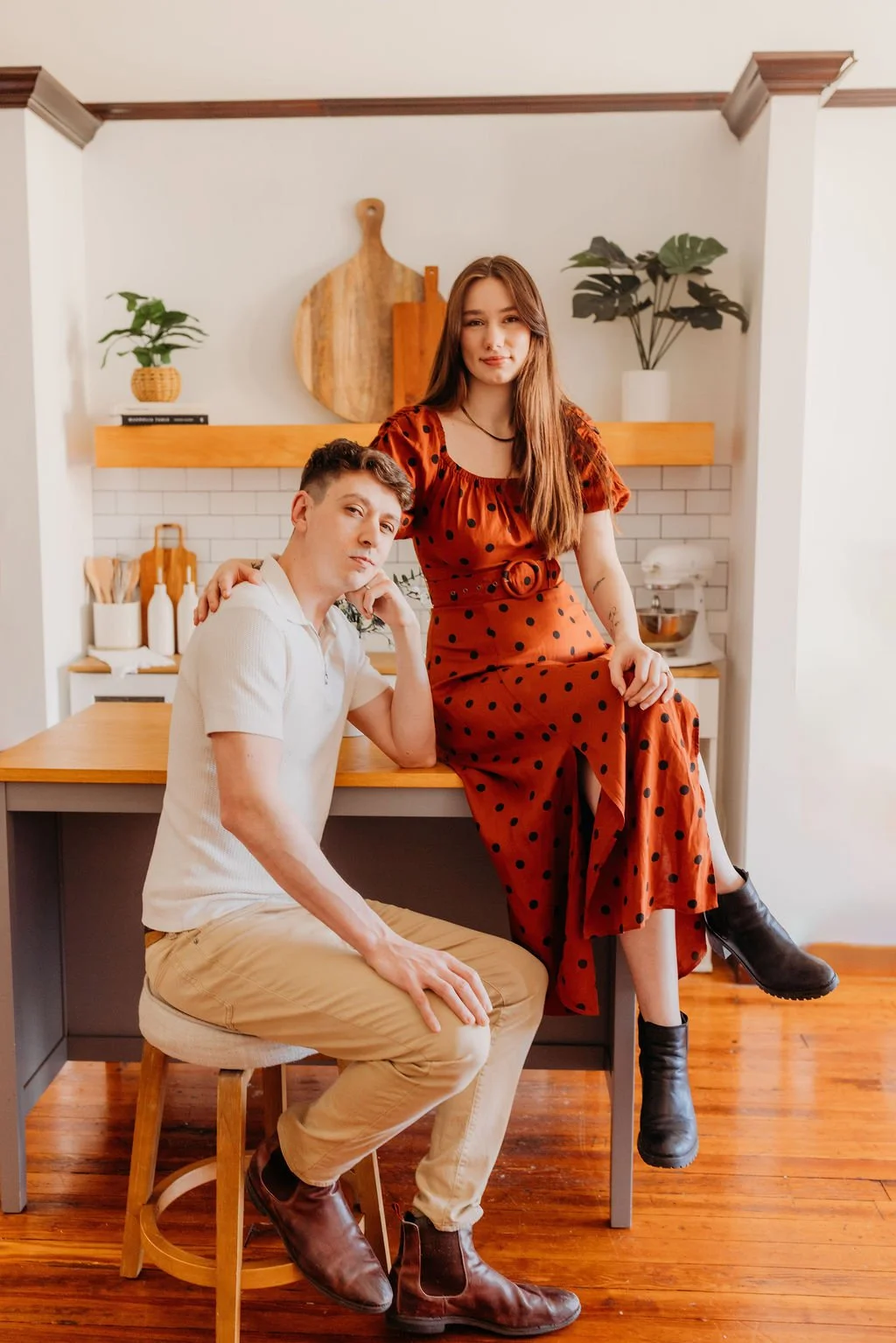 A young man and woman pose in a modern kitchen. The woman sits on the kitchen counter wearing a red polka dot dress and black boots. The man sits on a stool, wearing a white shirt and beige pants. The kitchen features white brick backsplash, wooden shelves, and minimal decor including plants and kitchen utensils.