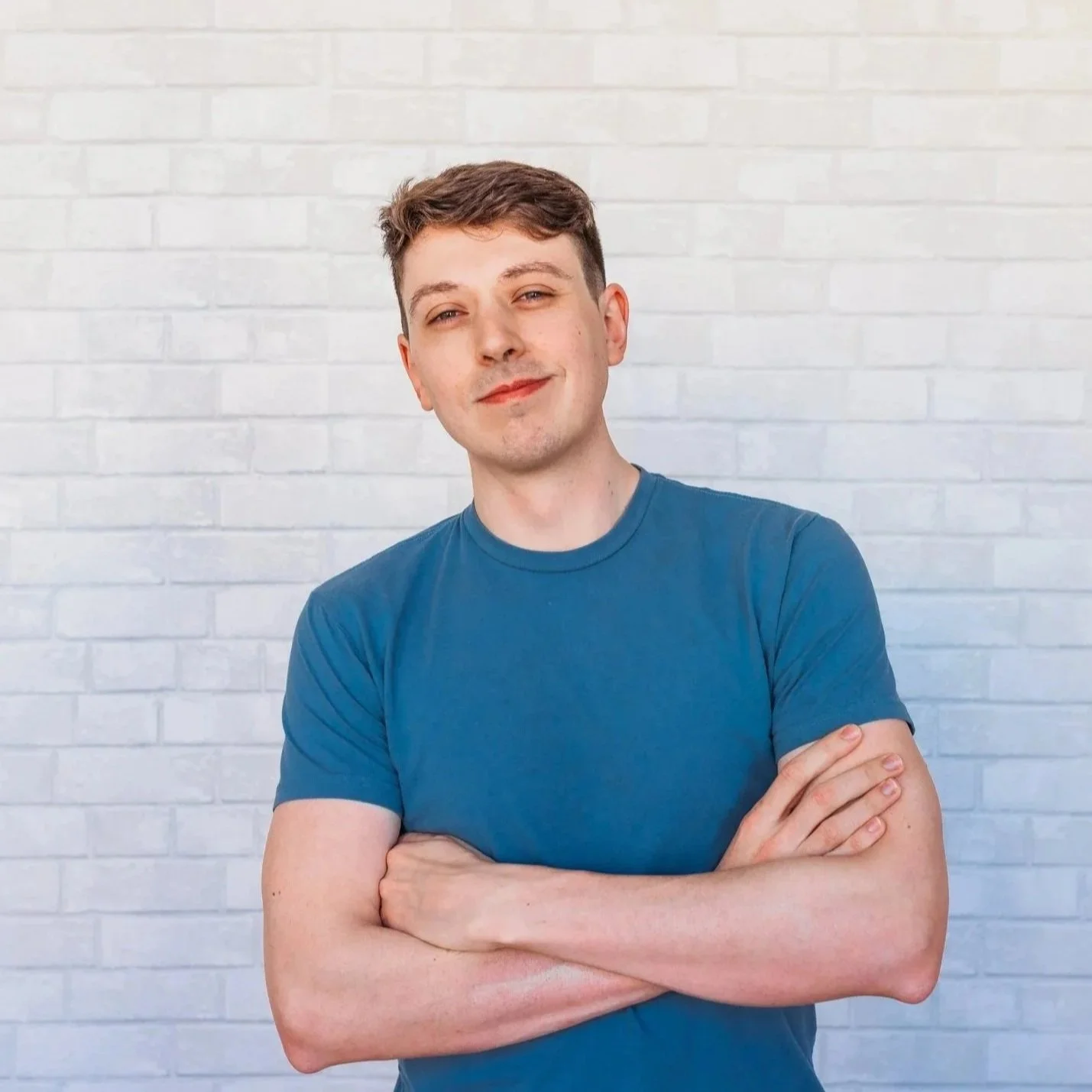 A young man with short brown hair and light skin, wearing a blue t-shirt, stands with arms crossed, smiling slightly, in front of a white brick wall.