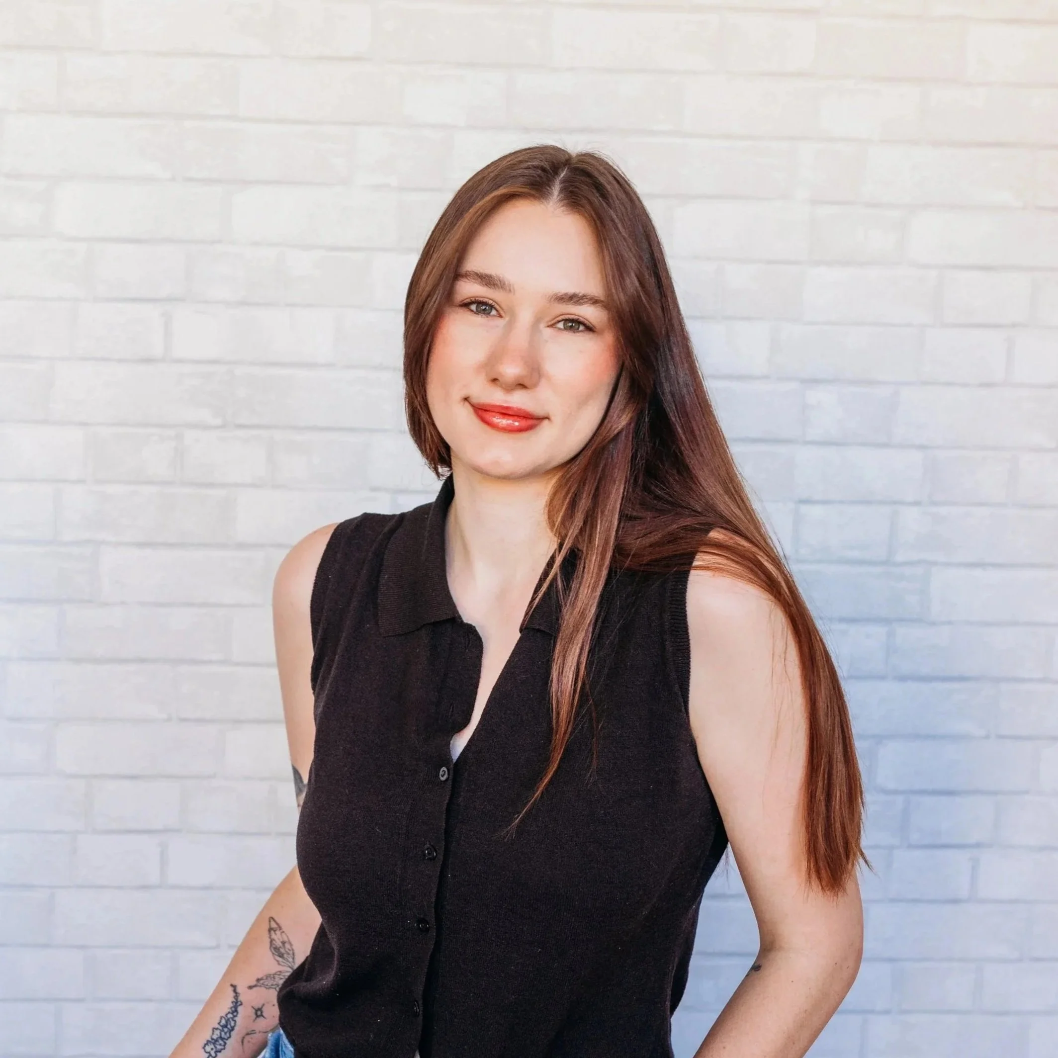 Young woman with long reddish-brown hair, wearing a sleeveless black top, smiling, standing in front of a white brick wall.