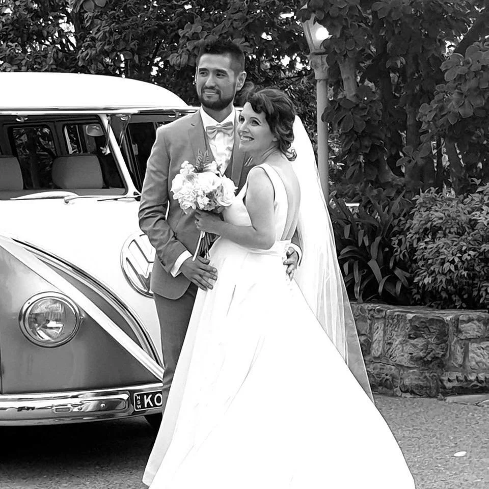 A bride and groom in wedding attire standing next to a vintage Volkswagen van, smiling and holding a bouquet of flowers in an outdoor setting with trees and bushes.