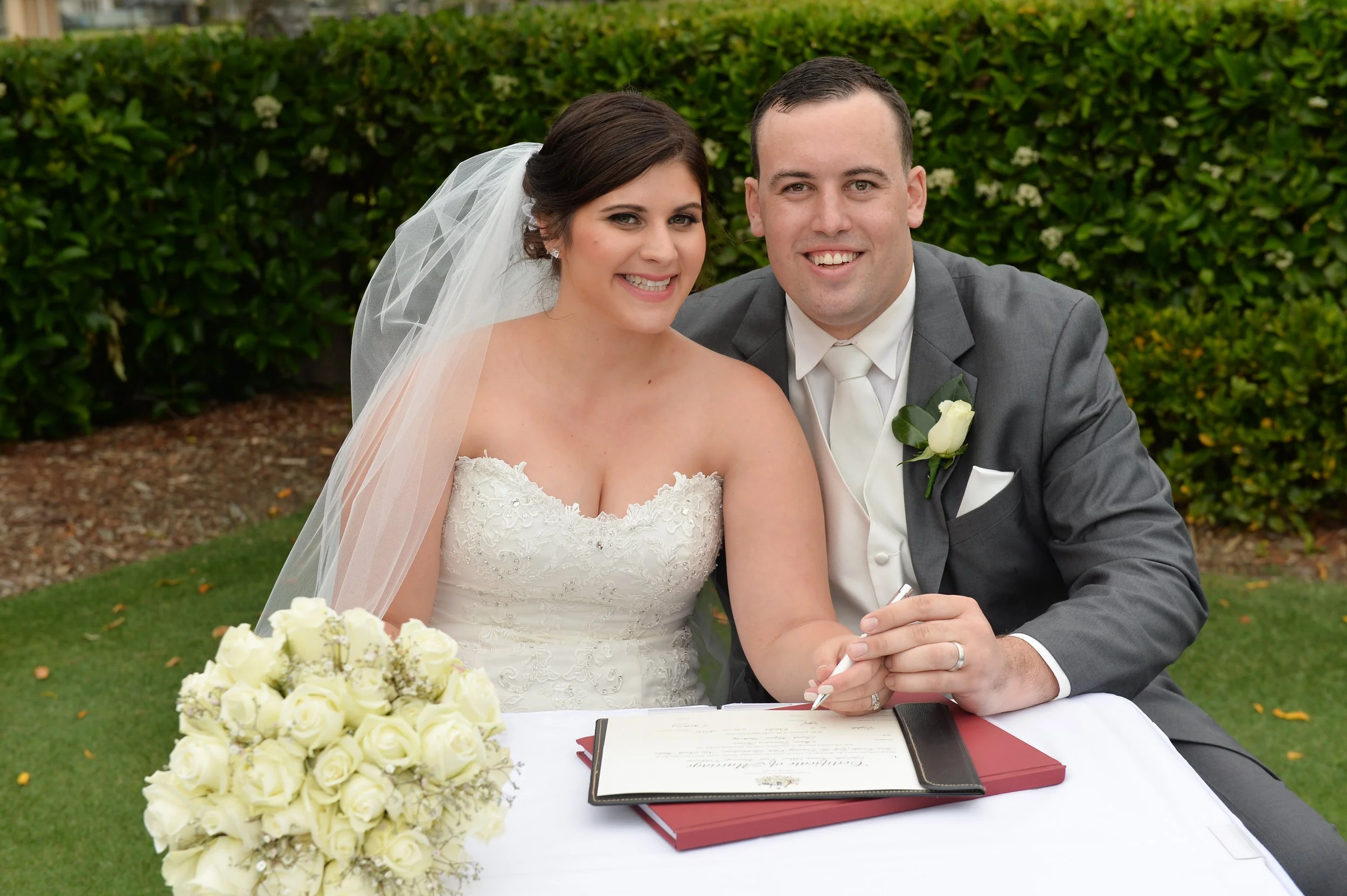 Bride and groom signing marriage certificate outdoors with green bushes in background.