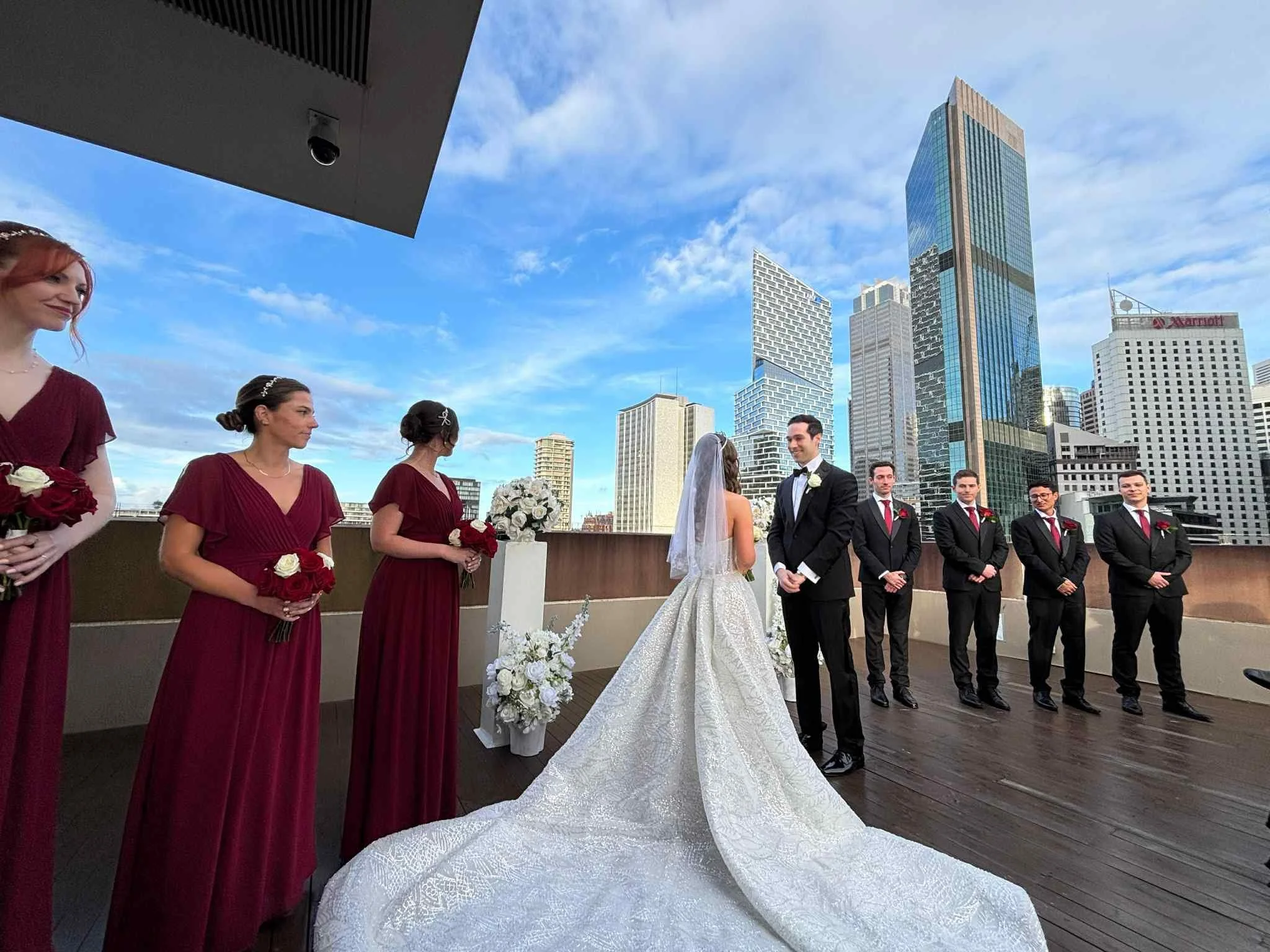 Wedding ceremony on a rooftop with a bride and groom facing each other, surrounded by bridesmaids in burgundy dresses holding red and white roses, and groomsmen in black suits with red ties, with a city skyline in the background under a partly cloudy