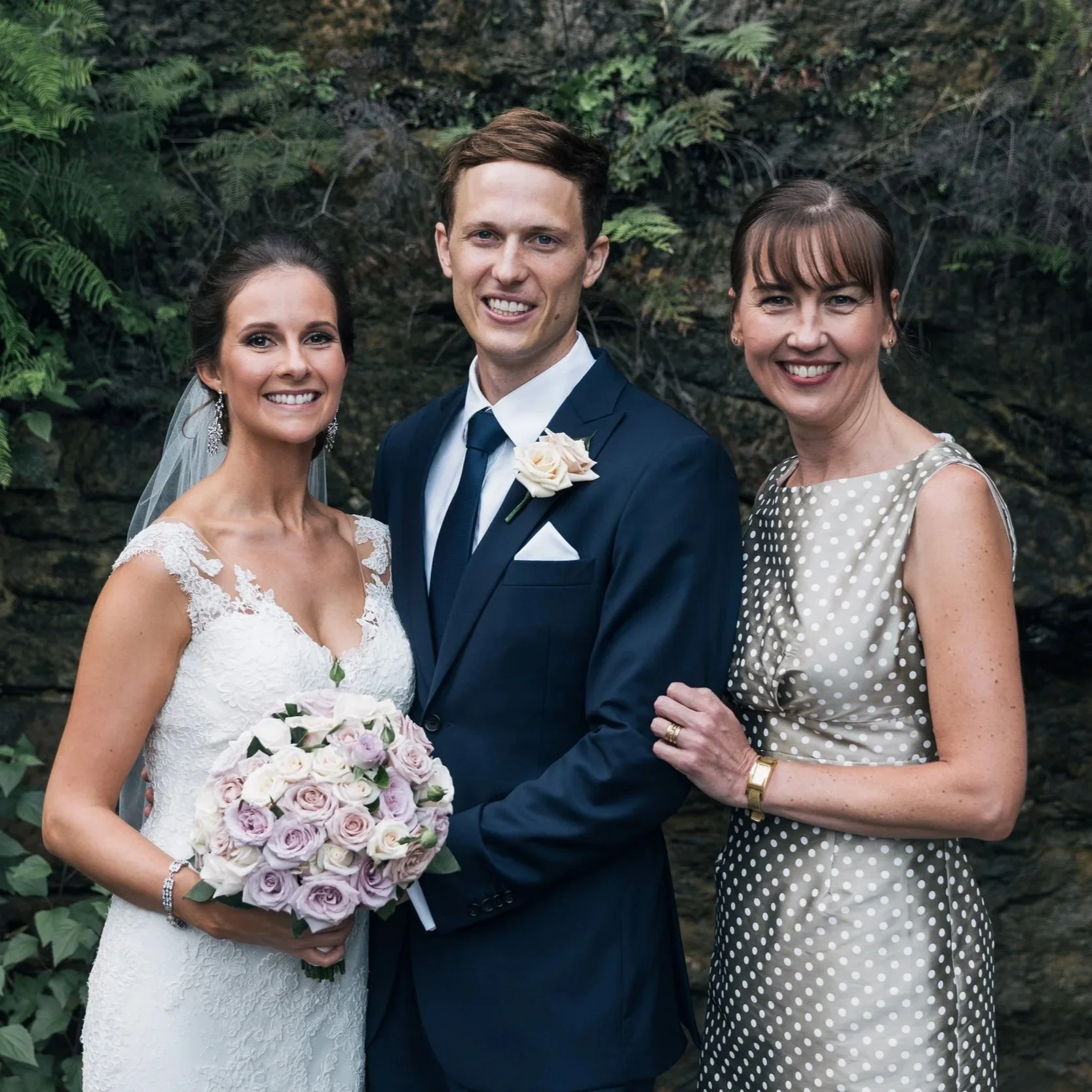 A bride in a white lace wedding dress holding a bouquet of light pink roses, standing next to a groom in a dark blue suit with a white dress shirt and a white flower boutonniere, accompanied by a woman in a polka dot dress, all smiling outdoors again