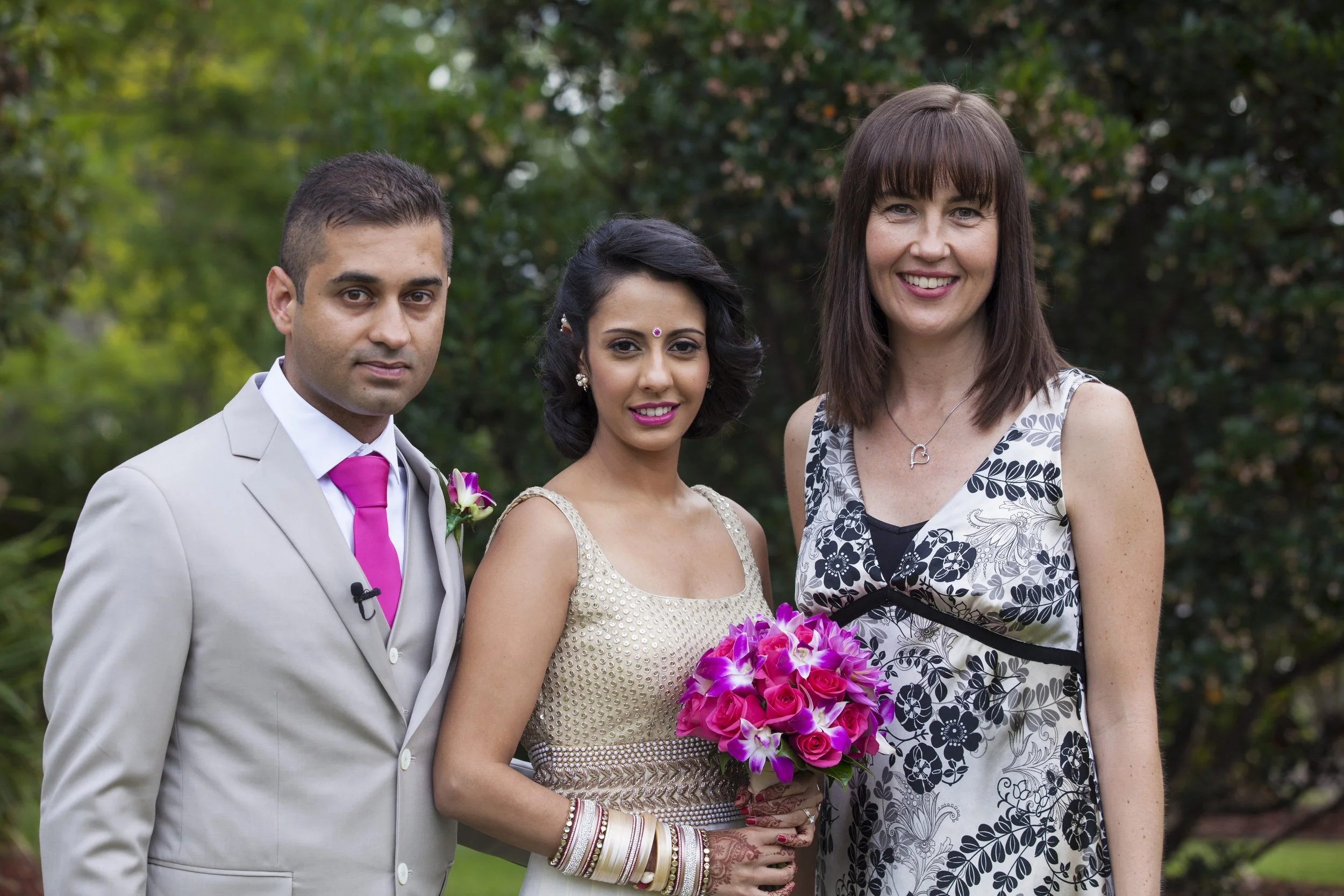 Three people standing outdoors, a man in a light gray suit with a pink tie, a woman holding a bouquet of pink and purple flowers, and another woman in a black and white floral dress, all smiling.