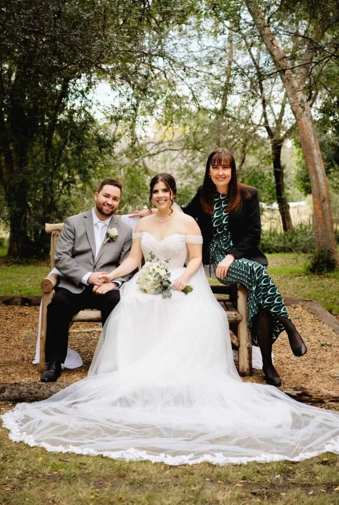 A bride in a white wedding dress with a bouquet, a groom in a gray suit, and a woman in a patterned dress and black blazer sitting on a wooden bench outdoors surrounded by trees.