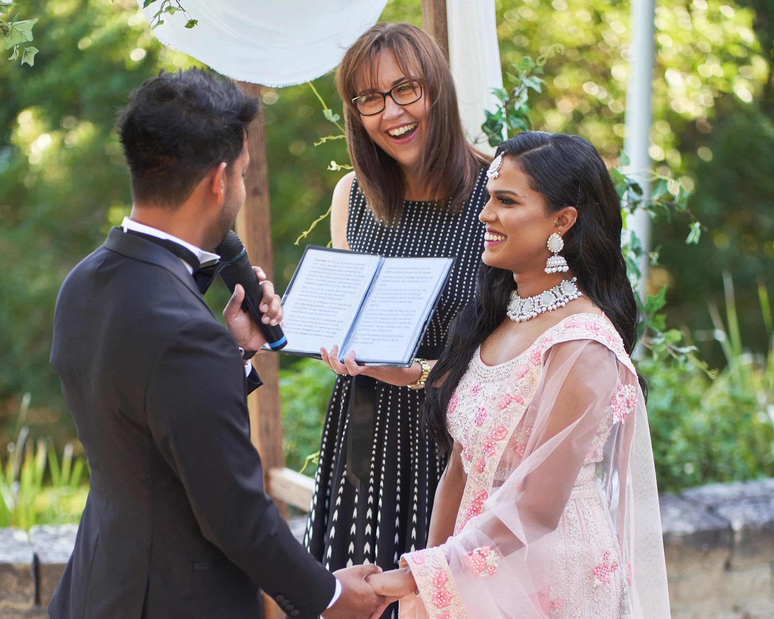 A wedding ceremony outdoors with a bride and groom holding hands, exchanging vows, with an officiant reading from a book, surrounded by greenery.