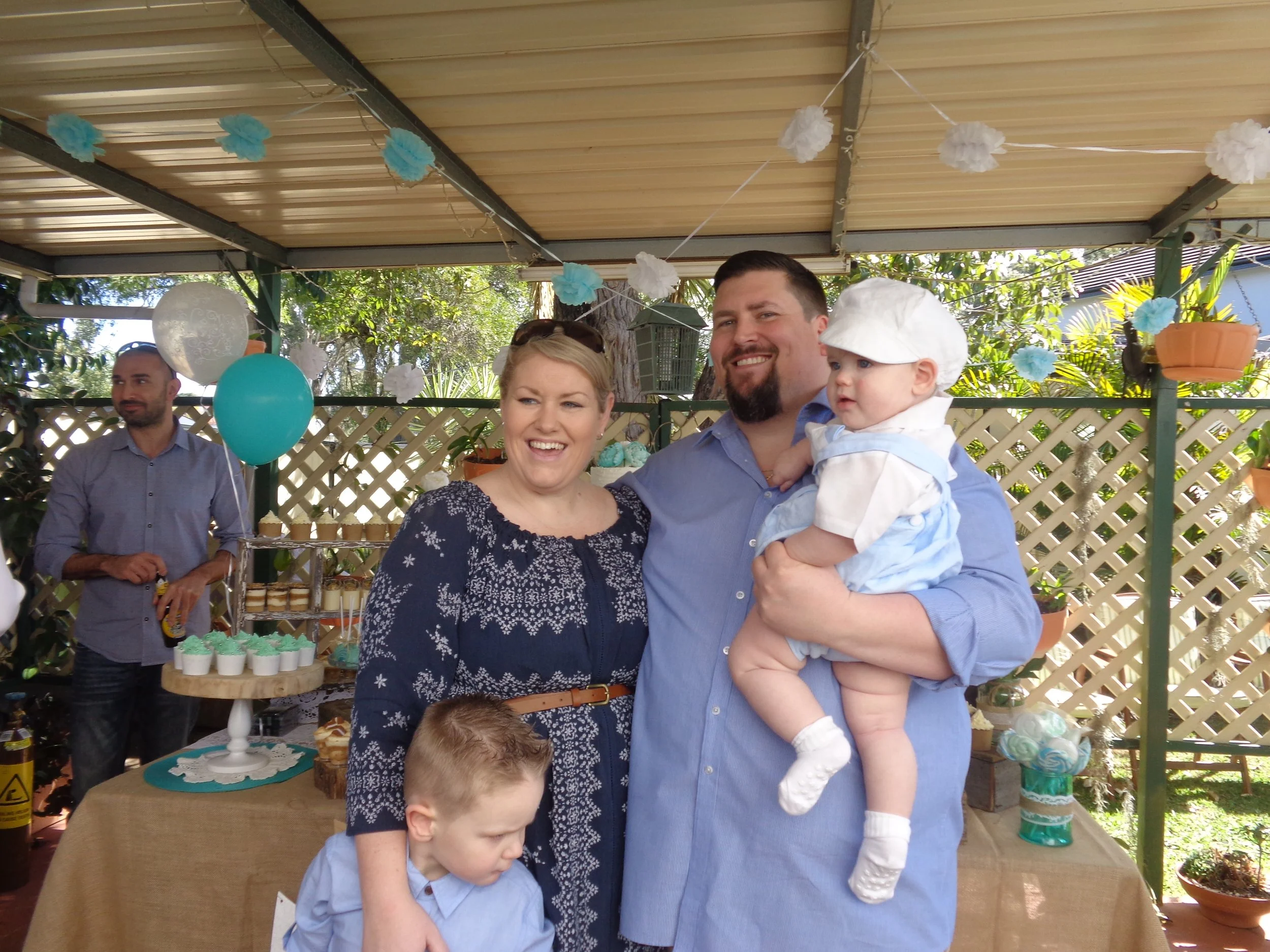 A family celebrating a child's birthday outdoors, with a woman, a man holding a small child dressed as a chef, and a young boy in front, smiling and celebrating under decorated blue and white balloons and paper pom-poms.
