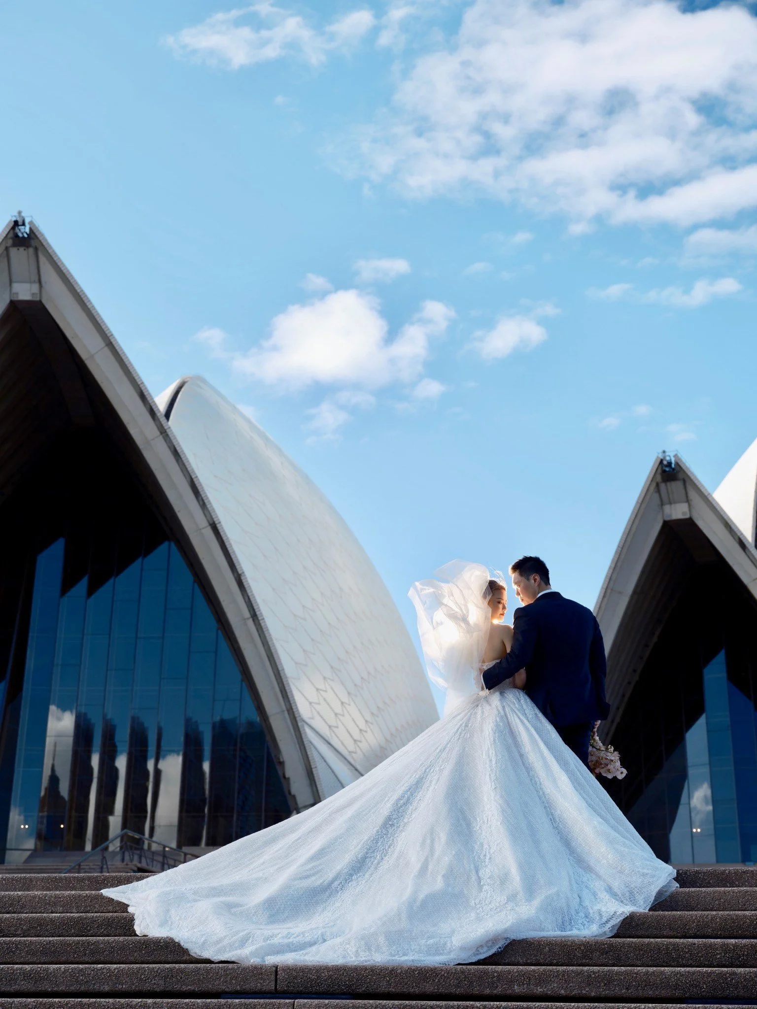 A bride and groom standing on steps outside Sydney Opera House. The bride wears a white wedding dress with a long train and veil, and the groom wears a dark suit. They are close to each other, with the bride holding a bouquet.